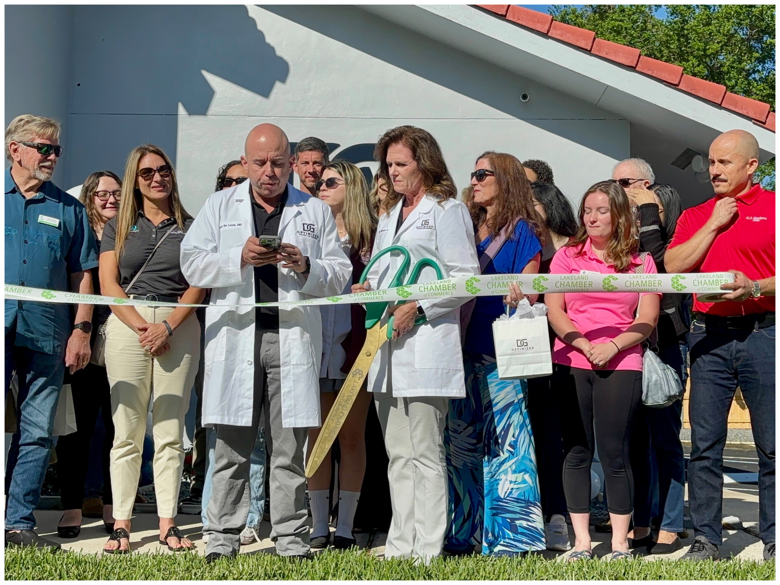 Group of people at a ribbon-cutting event, two in white lab coats holding large scissors.