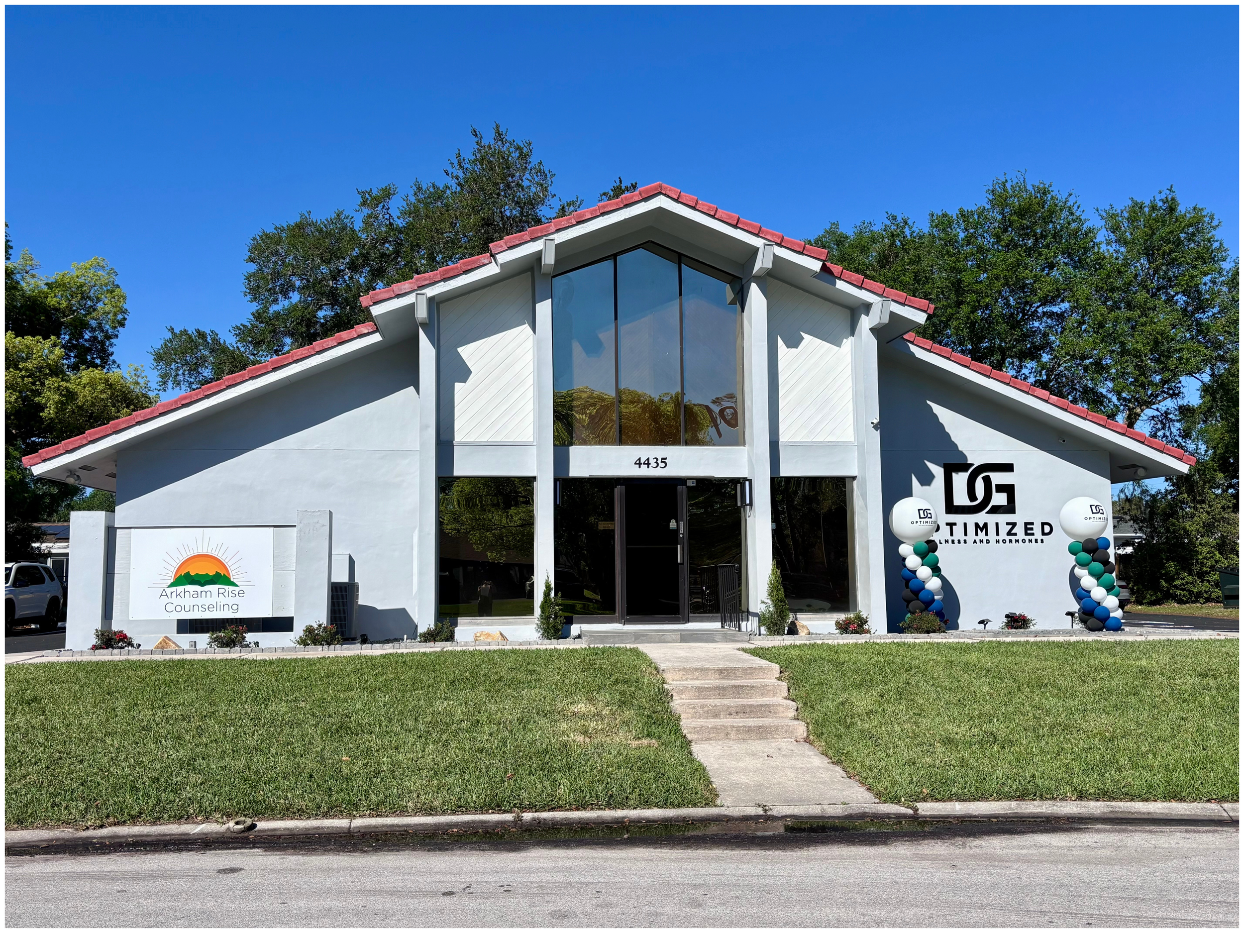 Front view of a modern building with a peaked roof, featuring signs for "Arkham Rise Counseling" and "Optimized Wellness and Hormones." The entrance is flanked by balloon columns. The building is white with a red roof and large windows. Trees and cle