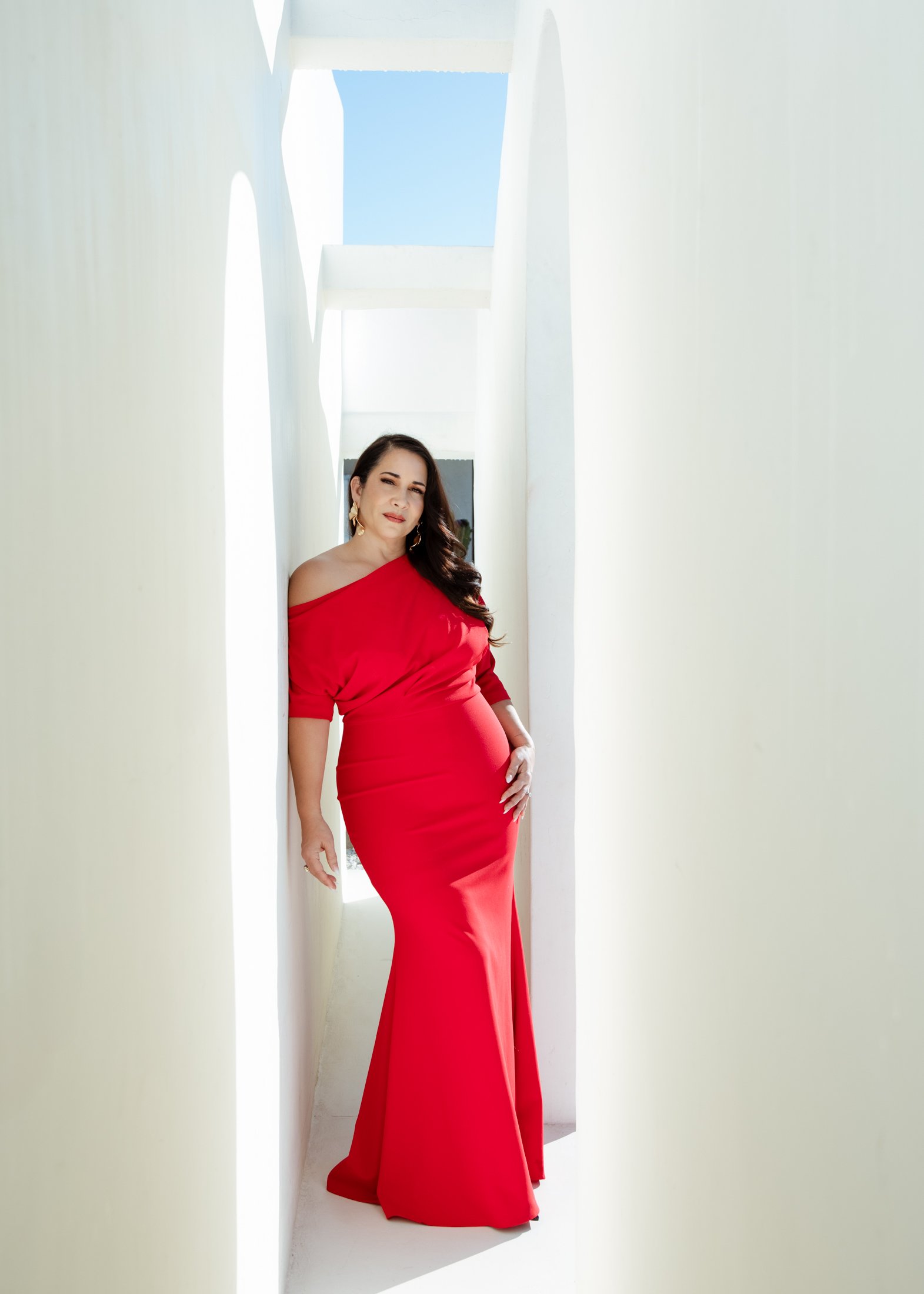 brunette woman standing against a white wall wearing a red gown 