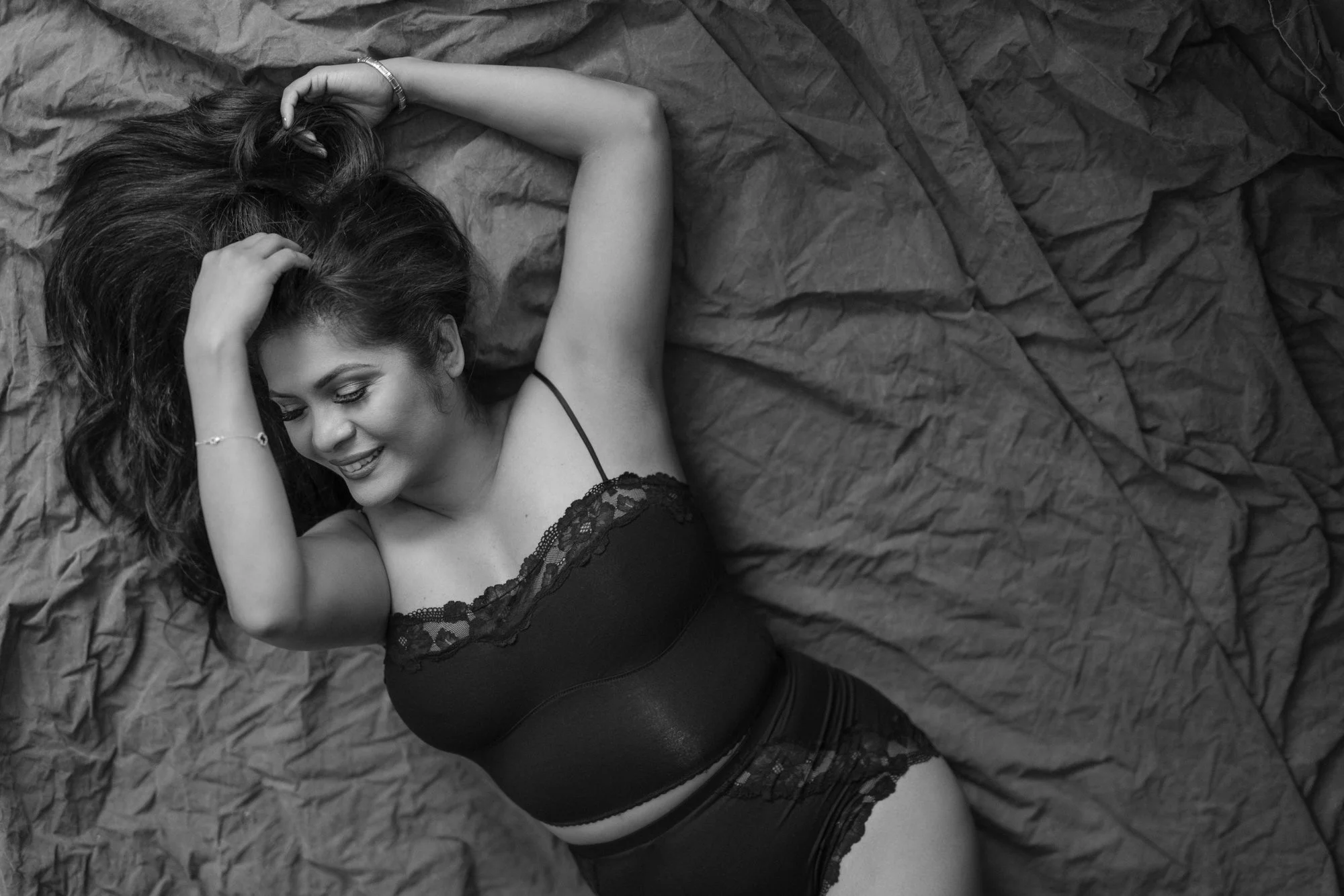 A woman lying on a bed with crinkled sheets, smiling and looking down. She has long dark hair, is wearing a black lace-trimmed top, and has one arm resting behind her head.