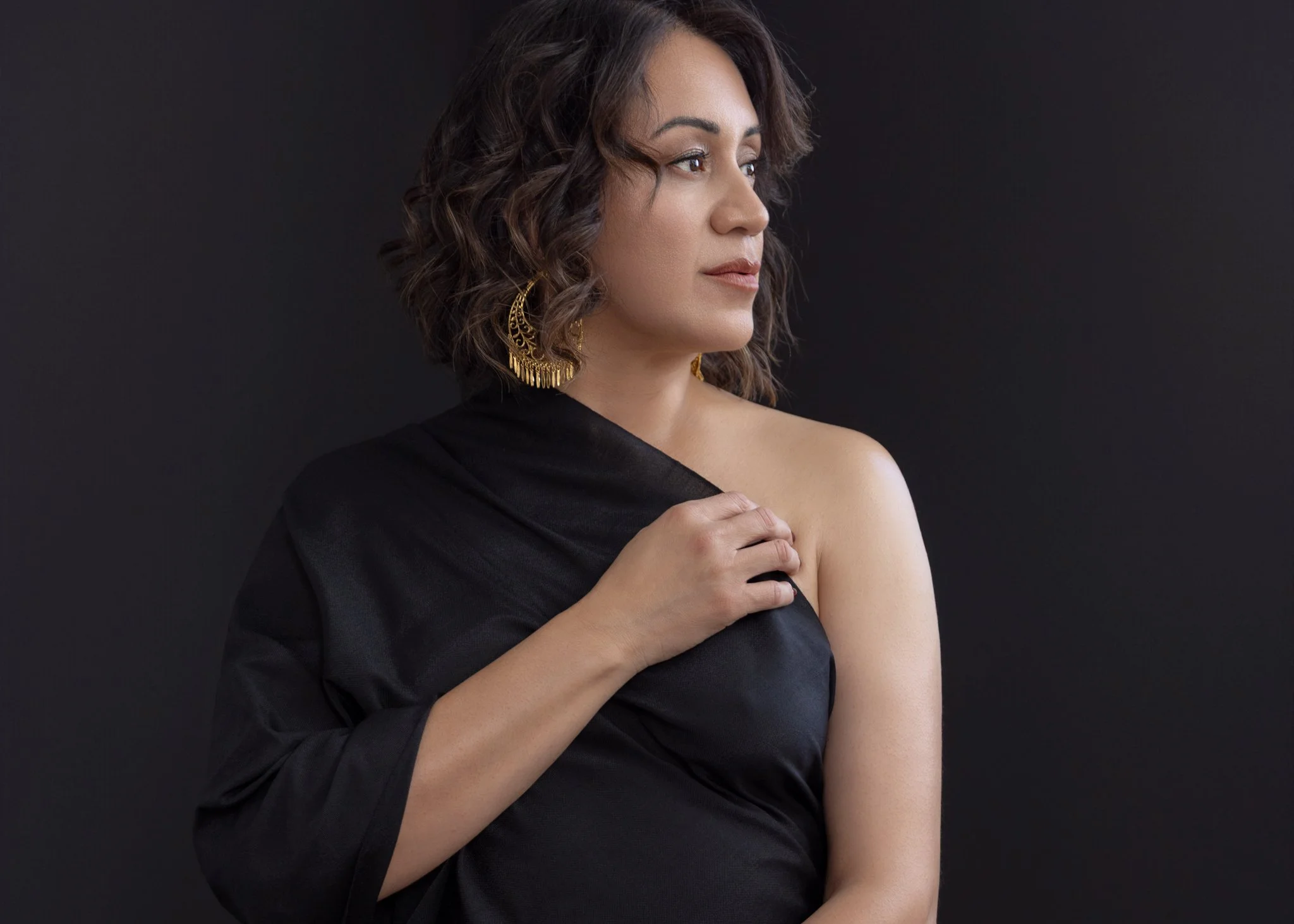 brunette woman wearing a black dress looking over her shoulder with gold earring dangling 