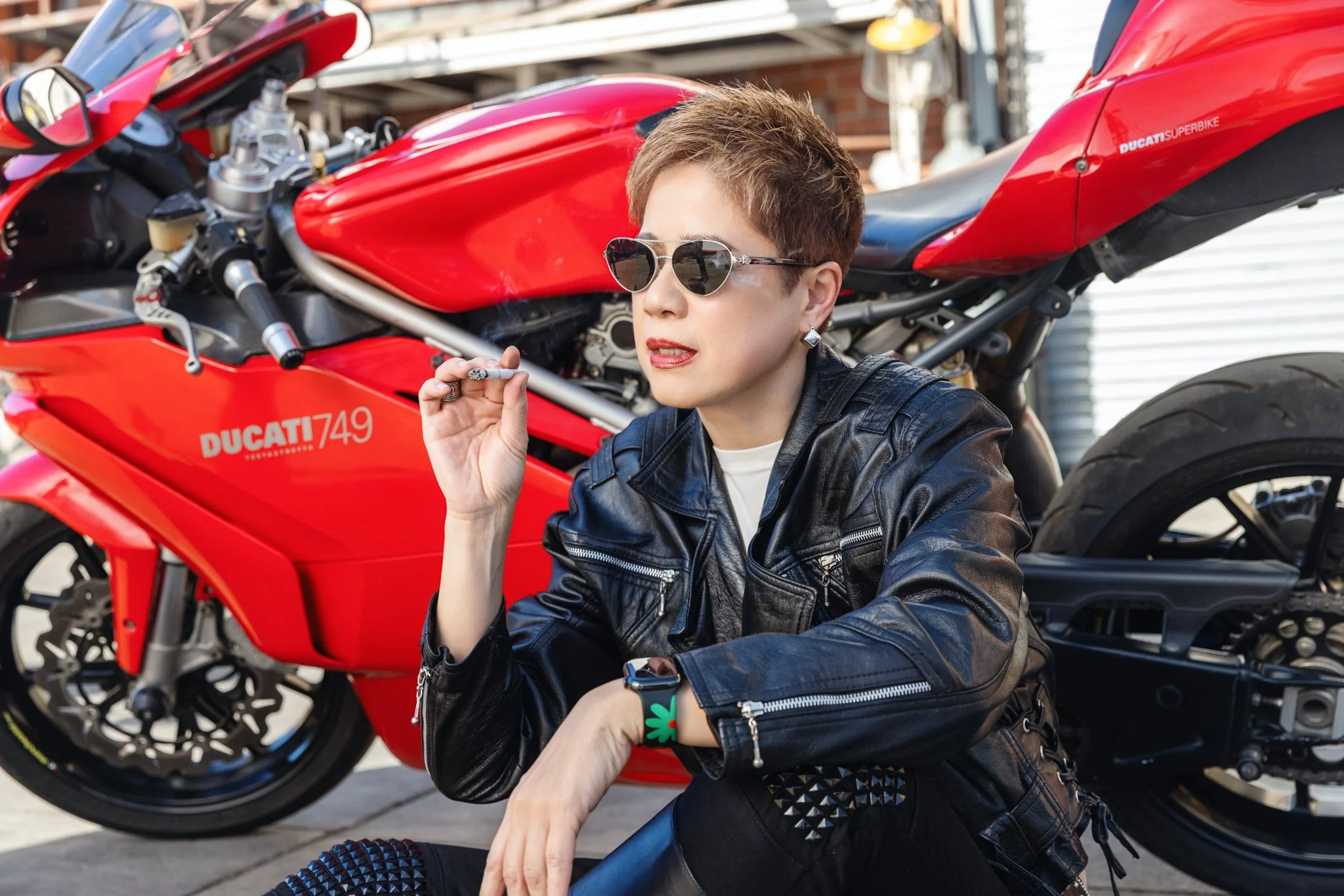 Woman wearing leather jacket smoking a cigarette sitting by a Ducati motorcycle