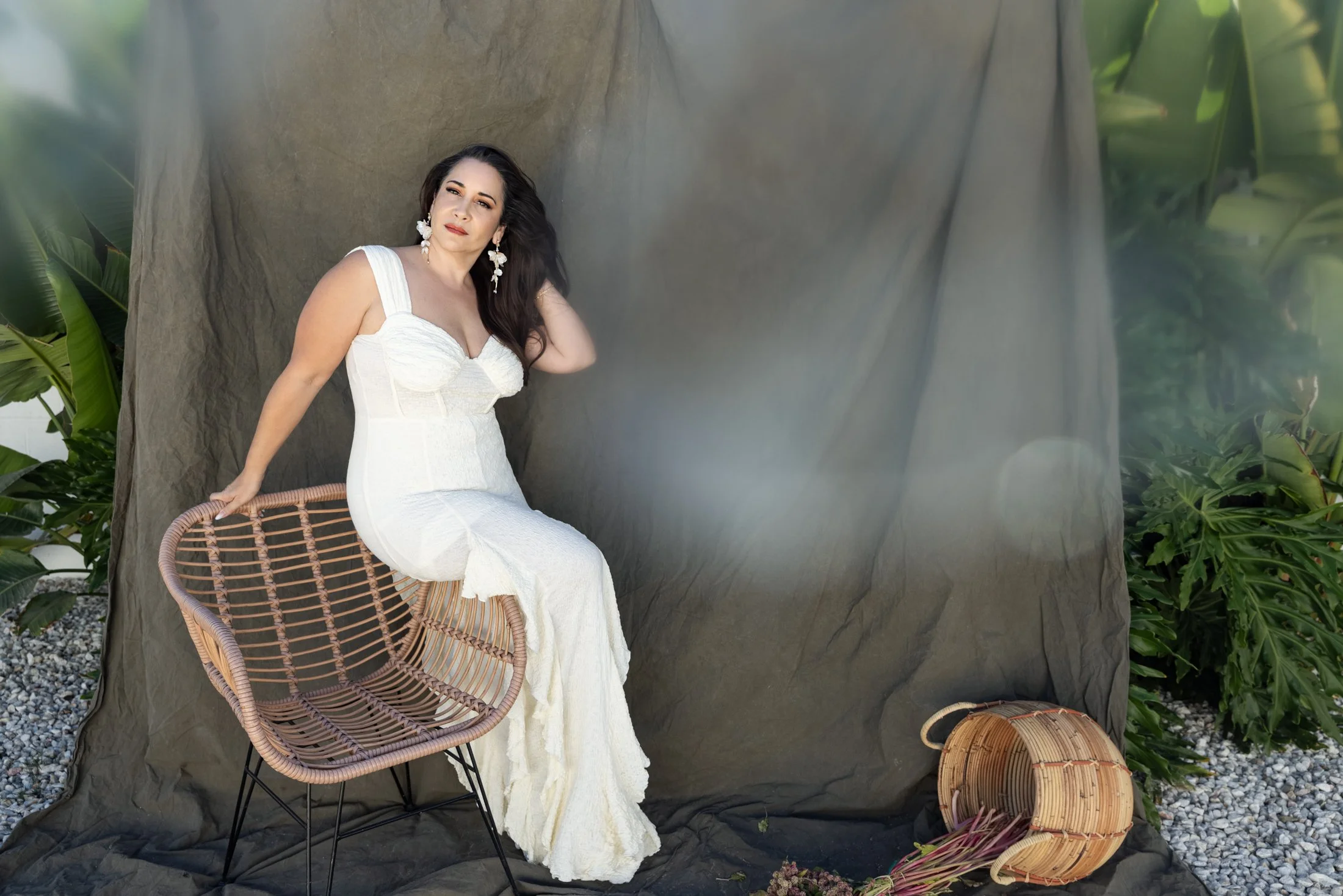 brunette woman wearing an ivory gown sitting in front of an olive green backdrop with lush greenery behind it 