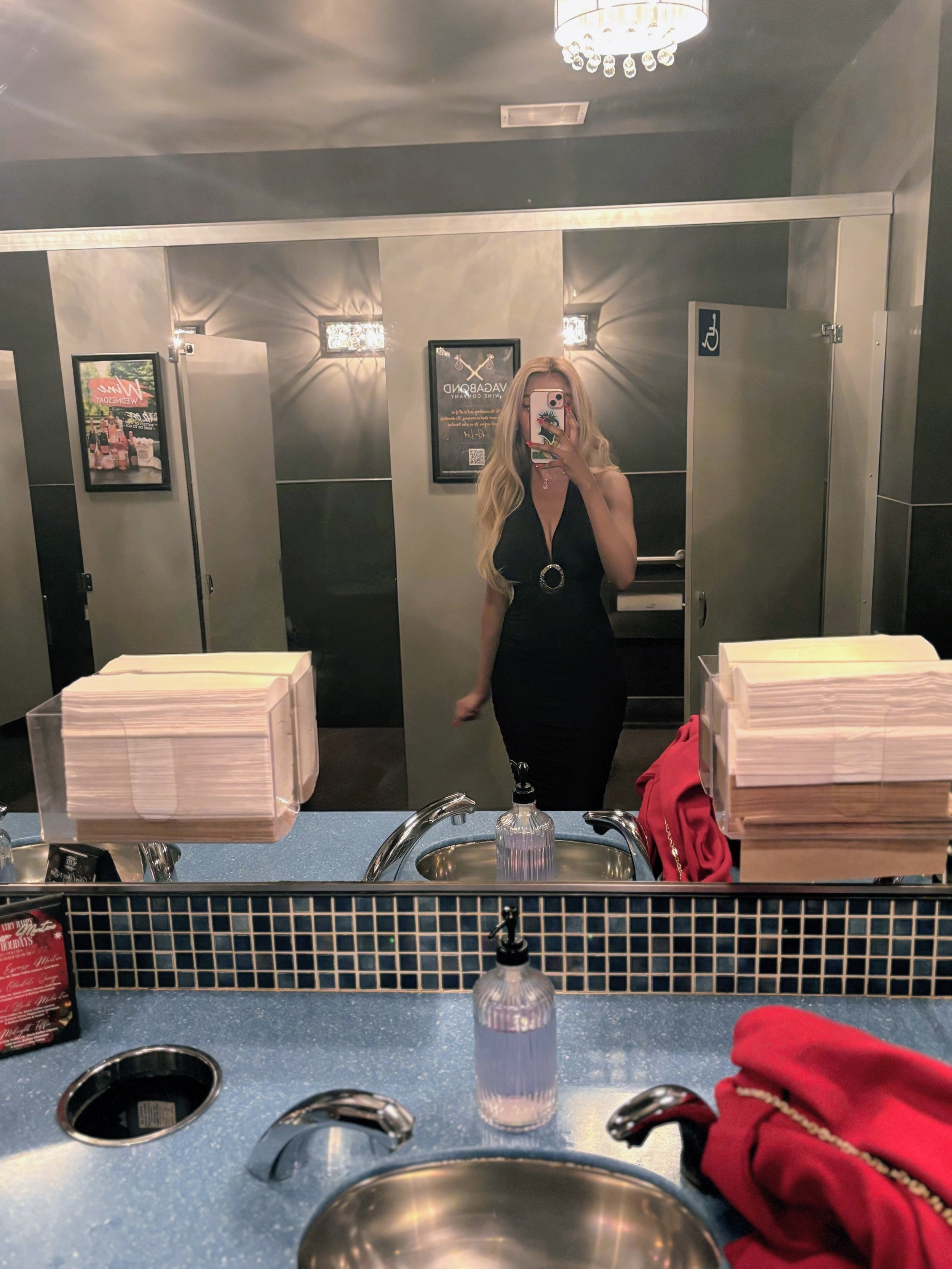 A woman taking a mirror selfie in a bathroom or restroom, with a curtain, framed pictures, and a drop ceiling with lights. She is wearing a black dress with a circular buckle and has long blonde hair.
