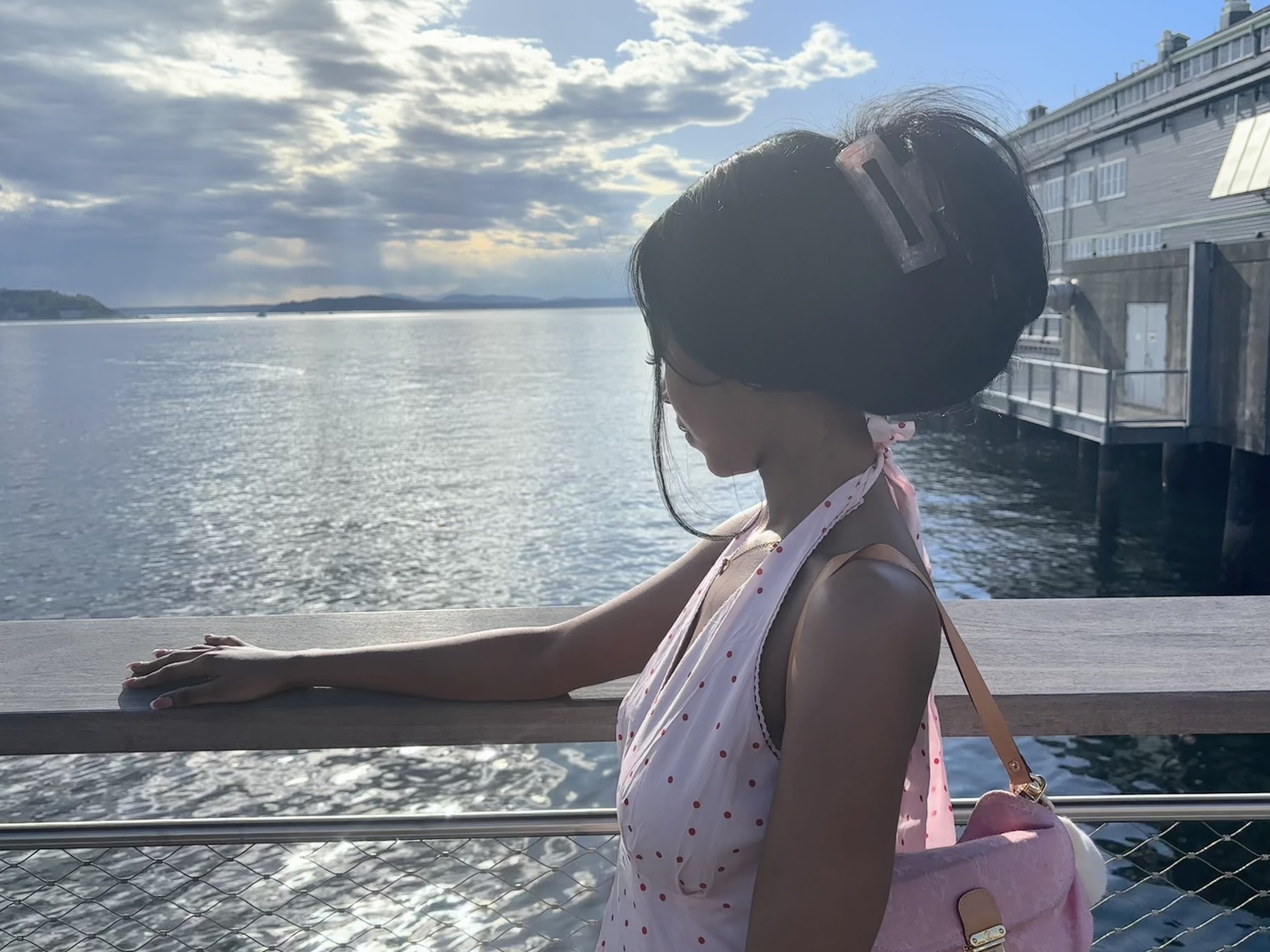 A young girl in a polka dot dress and pink bag leaning on a railing during the daytime near water with clouds in the sky.