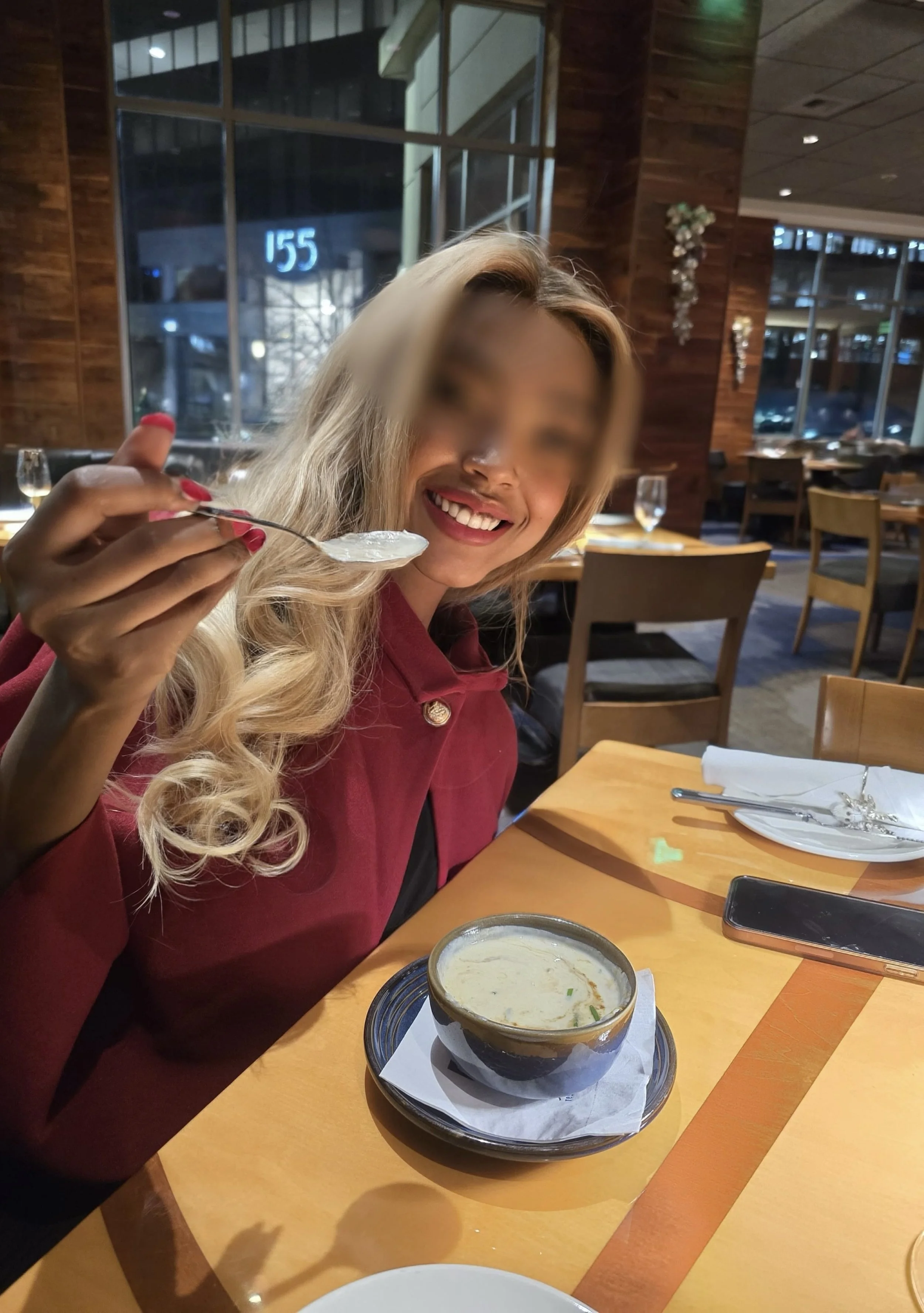 A smiling woman with long curly blonde hair, wearing a red or burgundy top, sitting at a restaurant table with a bowl of soup in front of her. She is holding a spoon with some soup. The restaurant has wooden walls, large windows, and decorative hangi