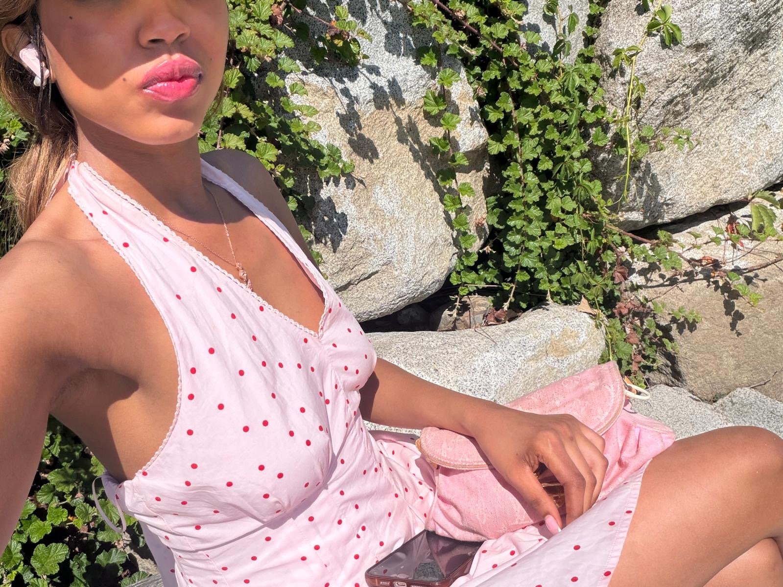 A woman sitting outdoors on large rocks and green plants, wearing a pink dress with red polka dots, pink purse, earrings, and lipstick, holding a phone and resting her hand on her lap.