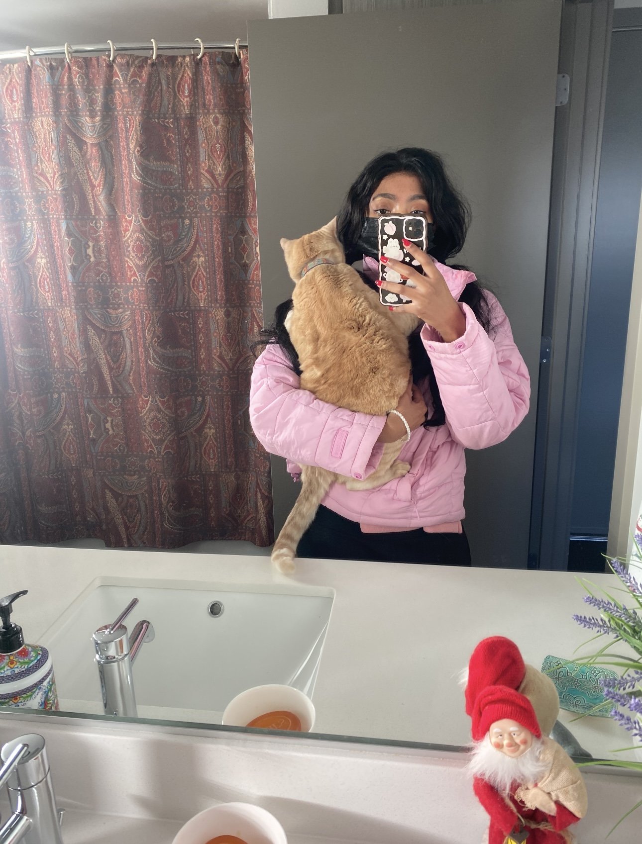 A woman with dark hair taking a mirror selfie in a bathroom, holding an orange tabby cat. She's wearing a pink jacket and a face mask. The bathroom has a paisley-patterned shower curtain, a white sink with soap dispensers, two bowls of orange liquid,