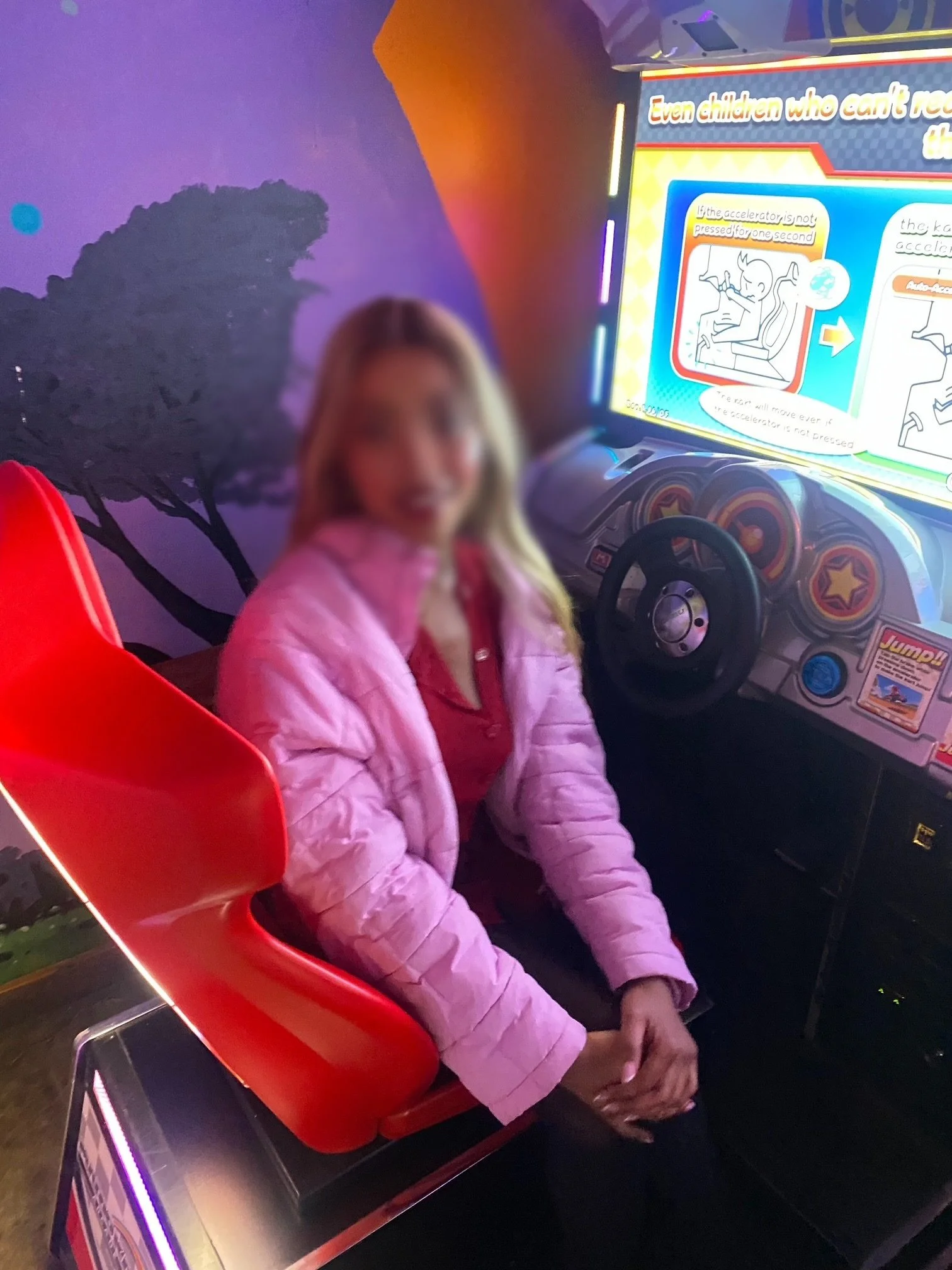 A woman seated in an arcade racing game seat with a pink puffer jacket and a red shirt, in front of a colorful game screen with instructions, in an arcade.