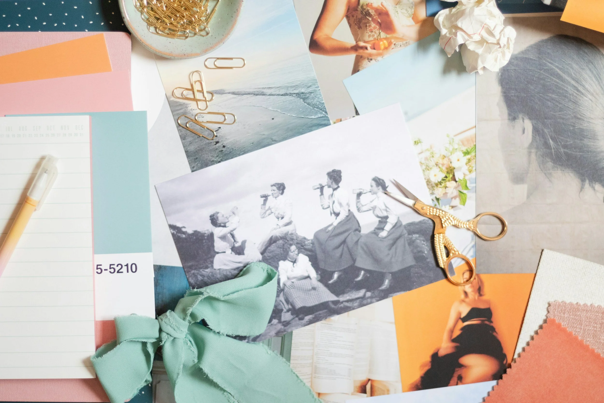 A cluttered desk with various photographs, fabric swatches, scissors, paper clips, a pen, and a ribbon, indicating an arts and crafts or sewing project.