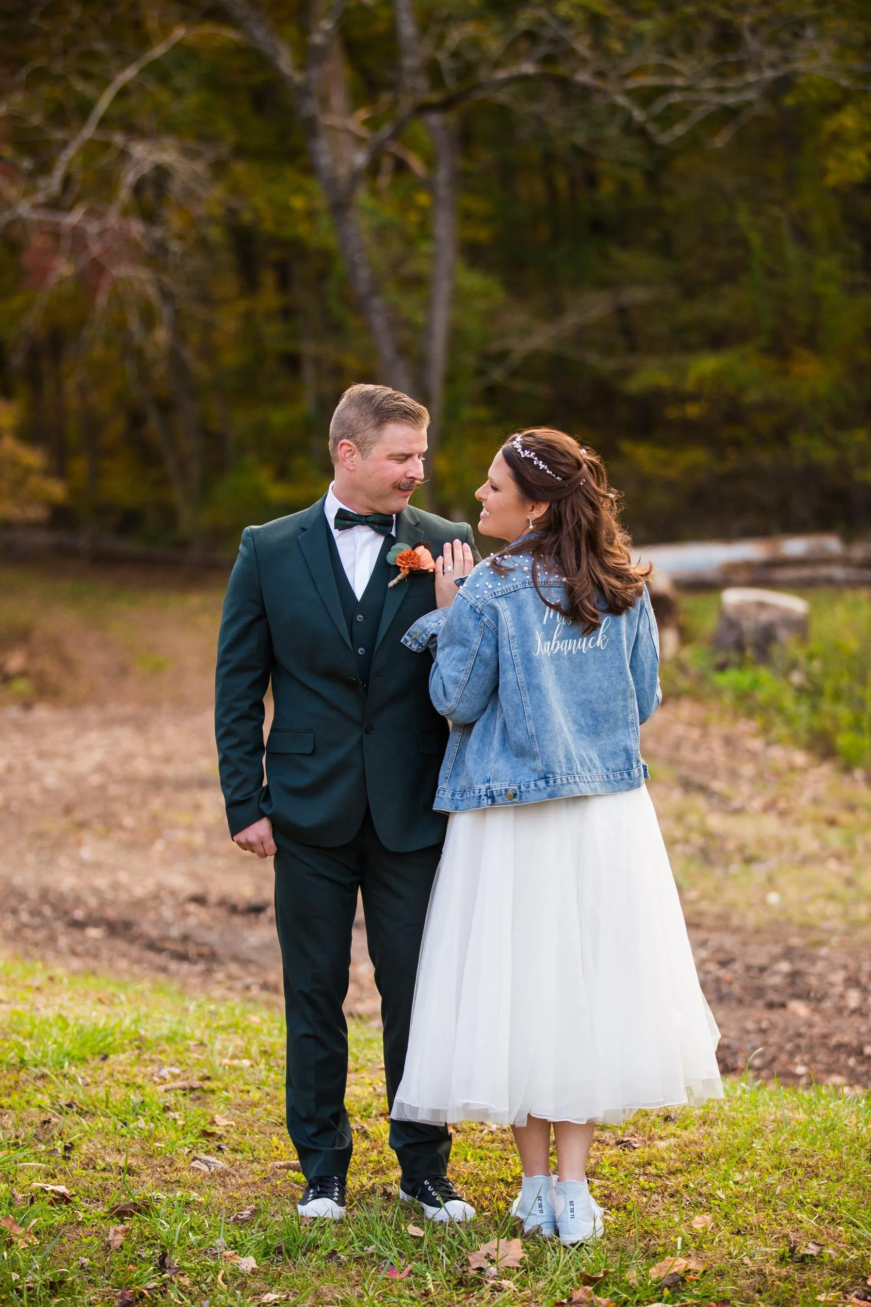 A couple, dressed in wedding attire, standing outdoors on grass with trees in the background during autumn. The man is wearing a dark suit with a bow tie, and the woman is wearing a white dress with a jean jacket. They are smiling and looking at each other.