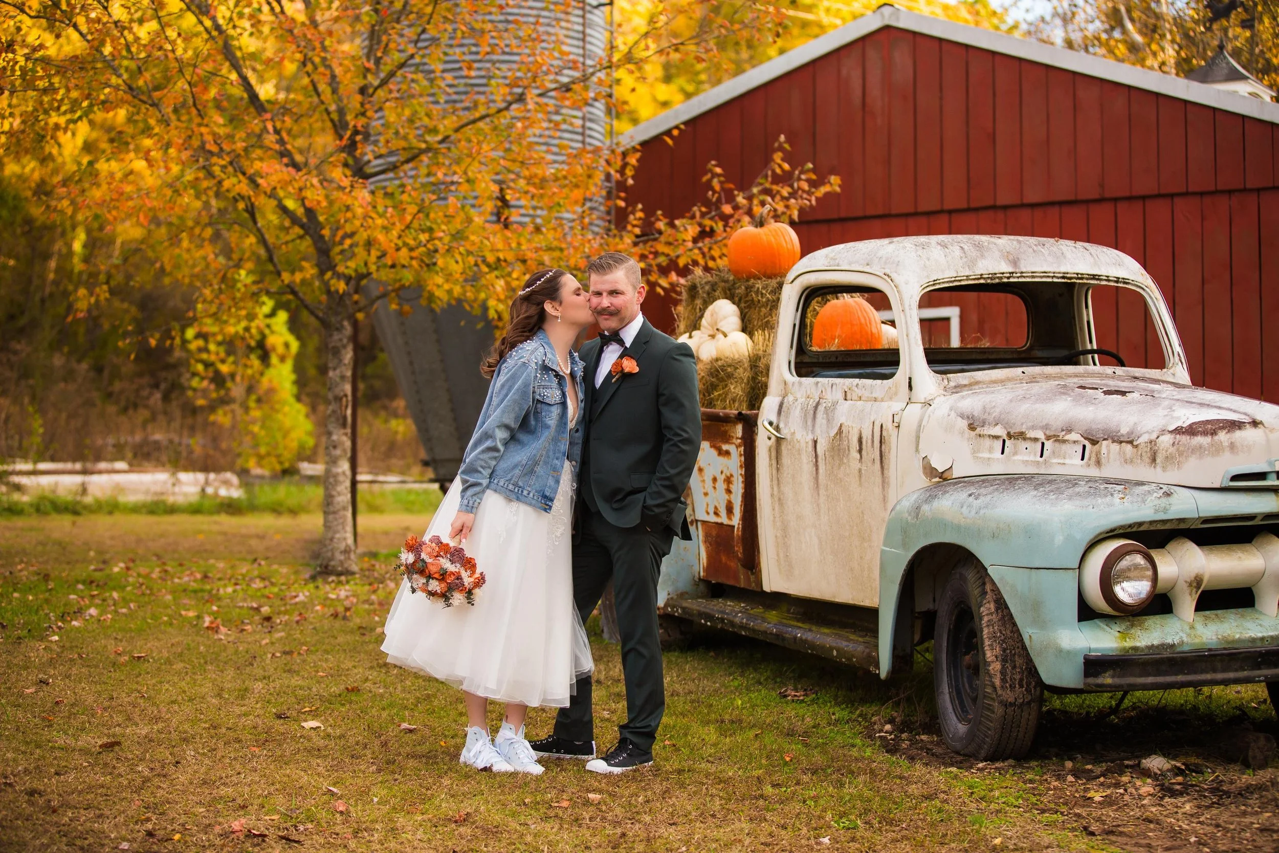 A bride and groom outdoors near an old, rusty pickup truck decorated with pumpkins and hay bales, with autumn trees and a red barn in the background.