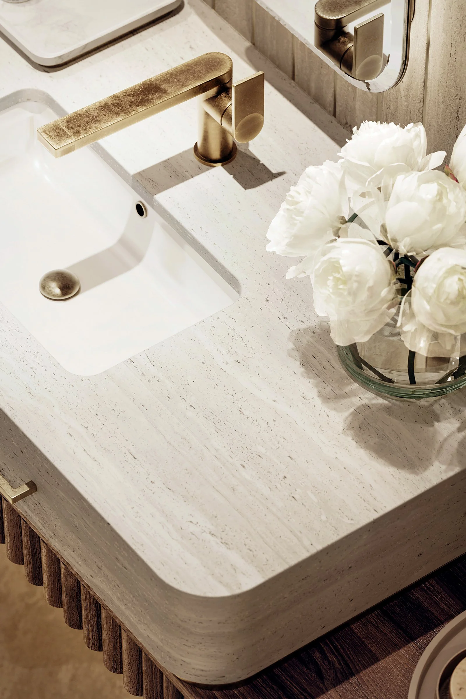 A close-up view of a modern kitchen sink with a matte gold faucet. The sink is embedded in a beige speckled countertop with a wood-textured cabinet below. A glass vase with white peony flowers is placed next to the sink, and a mirror is visible on the wall reflecting part of the faucet.
