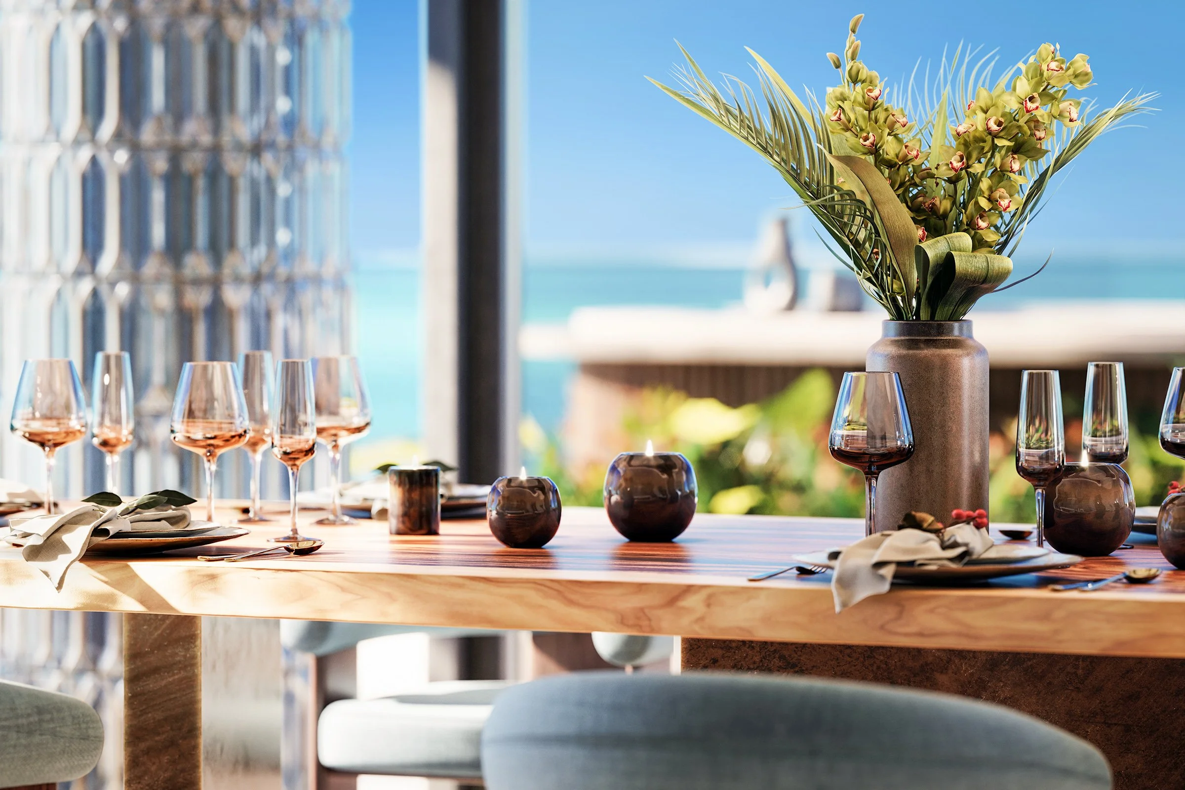 Elegant dining table setup with a centerpiece of green orchids and palm leaves in a gray vase, surrounded by wine glasses and candles, overlooking a beach with ocean and blue sky in the background.