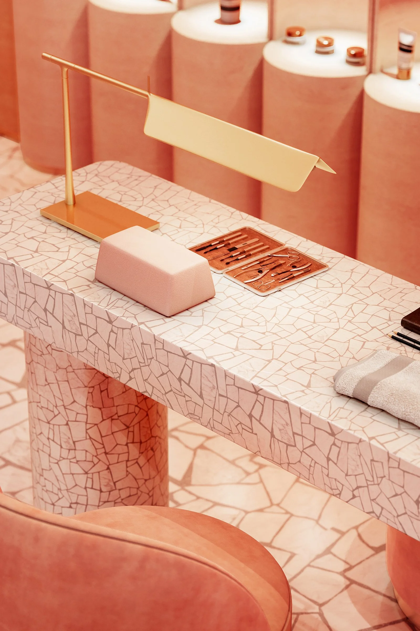 A manicure station with a beige cloth, a gold-colored lamp, and a set of manicure instruments on a mosaic table, with pink wallpapered walls and shelves displaying small bottles in the background.