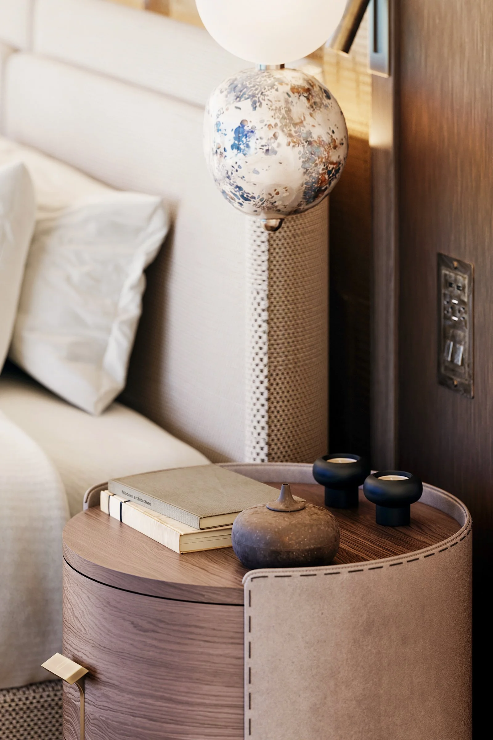 A bedroom scene featuring a bedside table with books and decorative objects, a bed with white bedding, a modern table lamp, and a wooden headboard with a wall outlet.