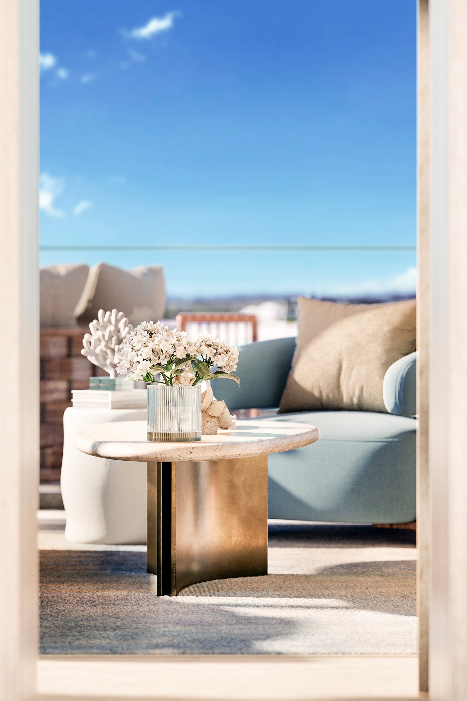 A round coffee table with a white vase holding white flowers, positioned in a cozy indoor space with cushions and a sofa, overlooking a bright outdoor balcony with a view of a clear blue sky and distant landscape.