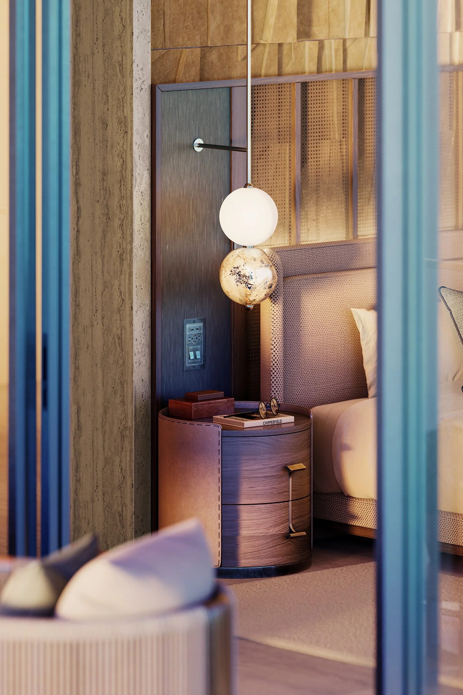 Interior of a modern hotel room with a nightstand, a hanging globe-shaped lamp, a patterned headboard, and a bed with pillows. The wall has wood paneling and an electrical outlet.
