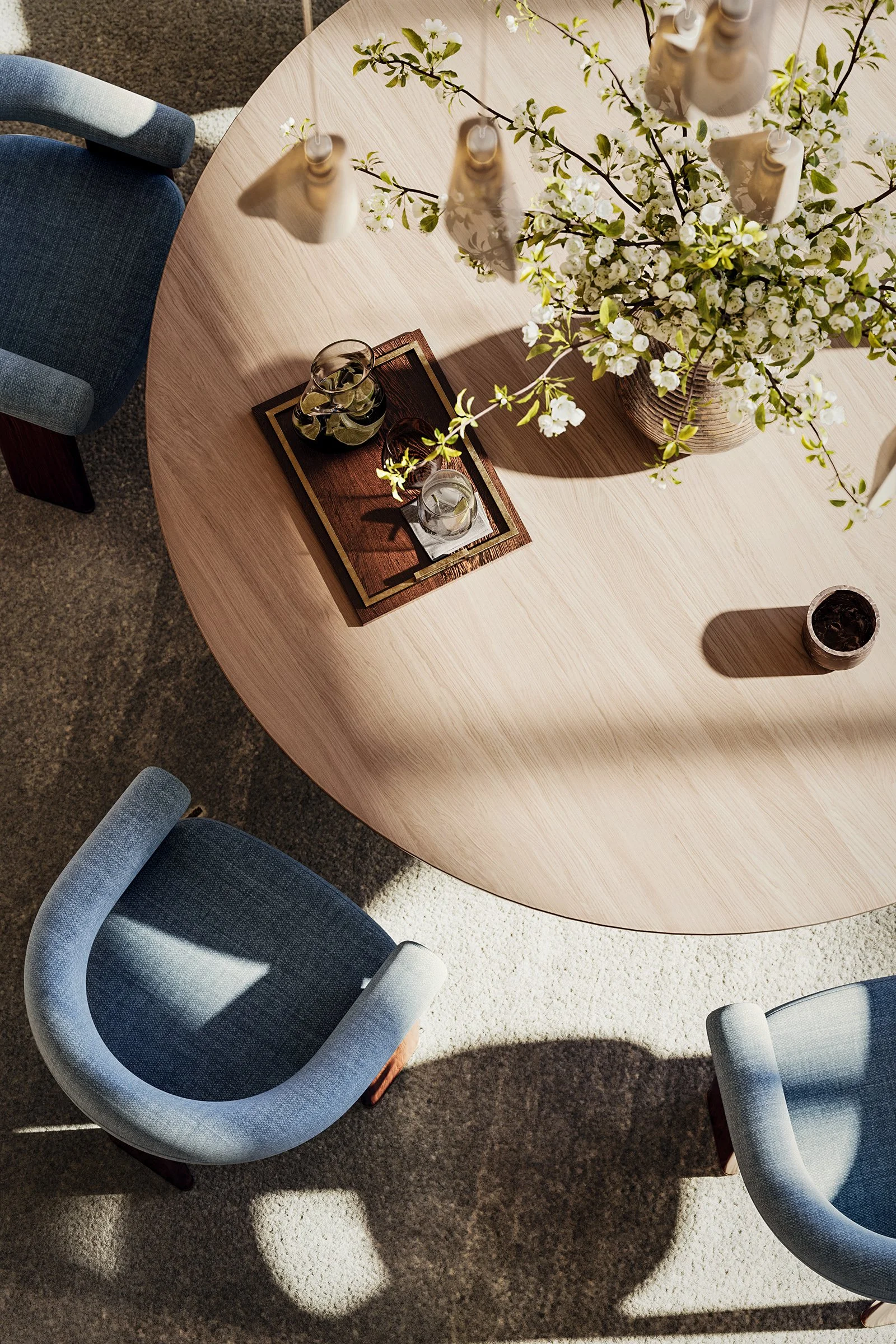 Top-down view of a wooden dining table with a large white floral arrangement in a woven basket and a tray with glasses and a pitcher of water. Two blue upholstered chairs are visible around the table. The table is partially in sunlight, casting shadows.