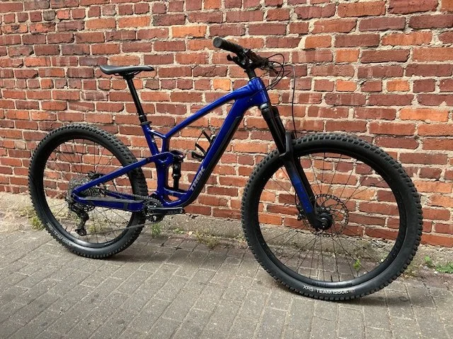 Blue mountain bike with front suspension and disc brakes, parked against a brick wall.
