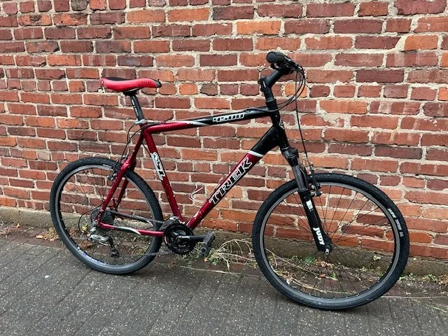 Red and black Trek mountain bike leaning against a red brick wall on a concrete sidewalk.