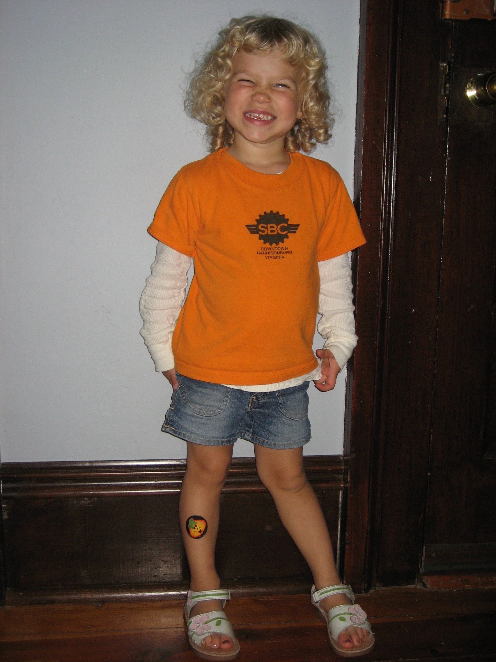 A young girl with curly blonde hair standing indoors against a white wall and wooden door frame, smiling with squinted eyes, wearing an orange T-shirt, denim shorts, white long-sleeve shirt underneath, and white sandals, with a lion sticker on her right leg.
