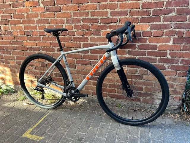 A gray Trek mountain bike with black tires and a front suspension fork, parked against a brick wall on a paved sidewalk.