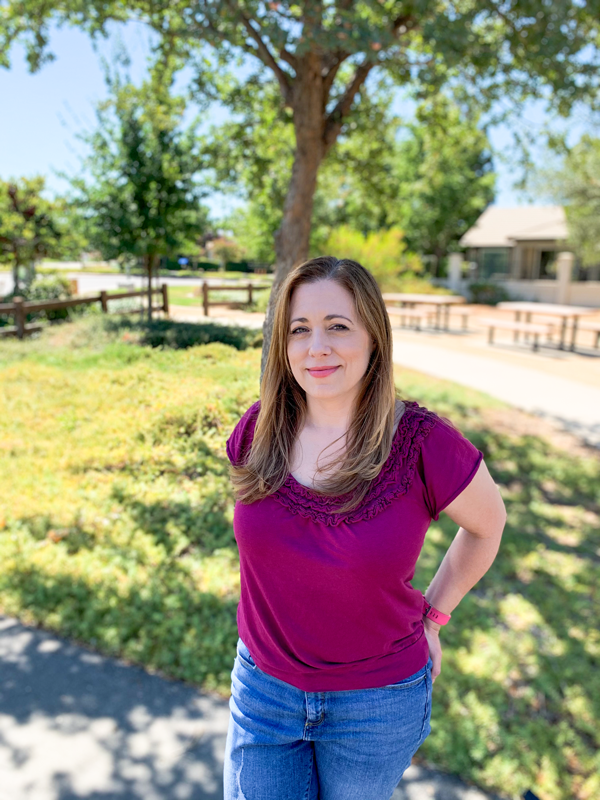 Beth Cardwell in a purple top and blue jeans standing outdoors in a park, with trees and a building in the background.