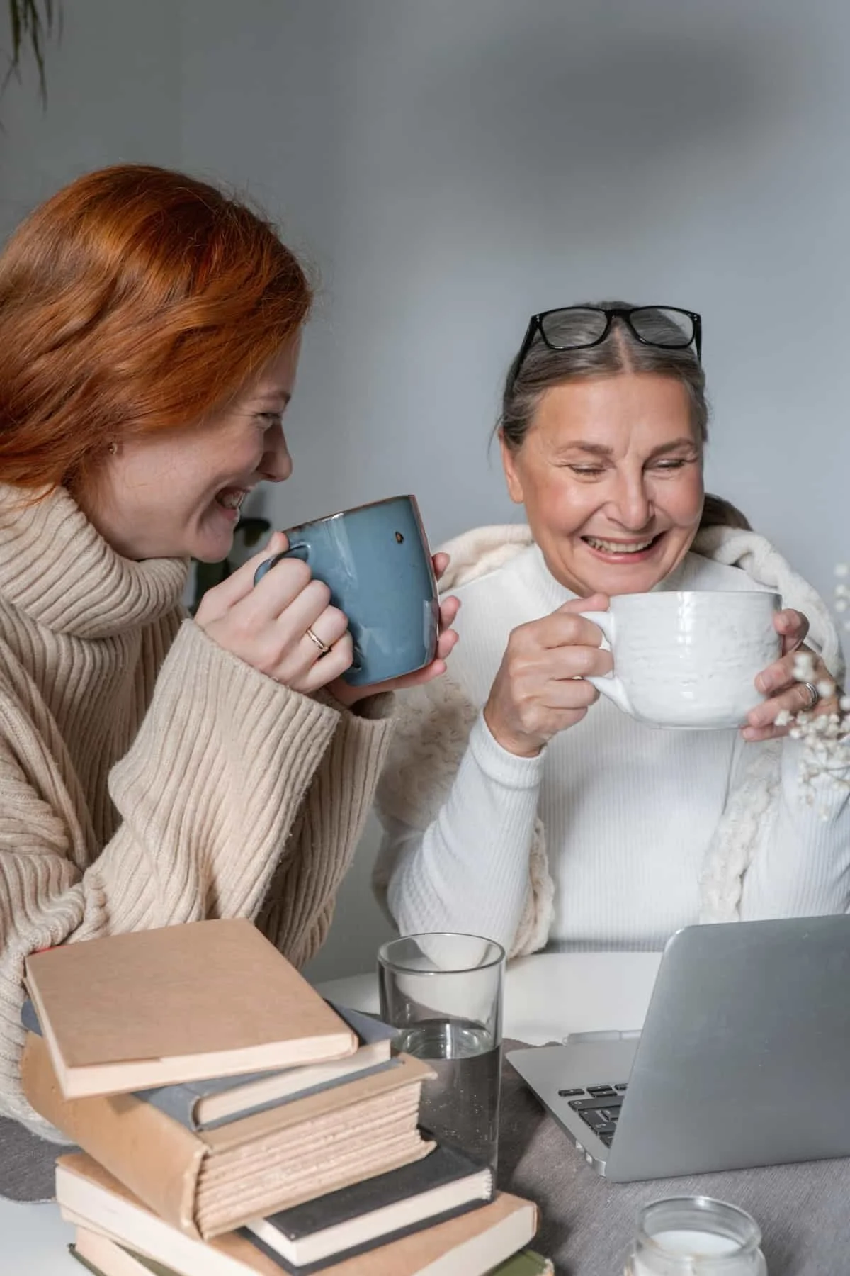 Two women enjoying coffee and tea while sharing a laugh at a table with books, a laptop, and glasses of water.