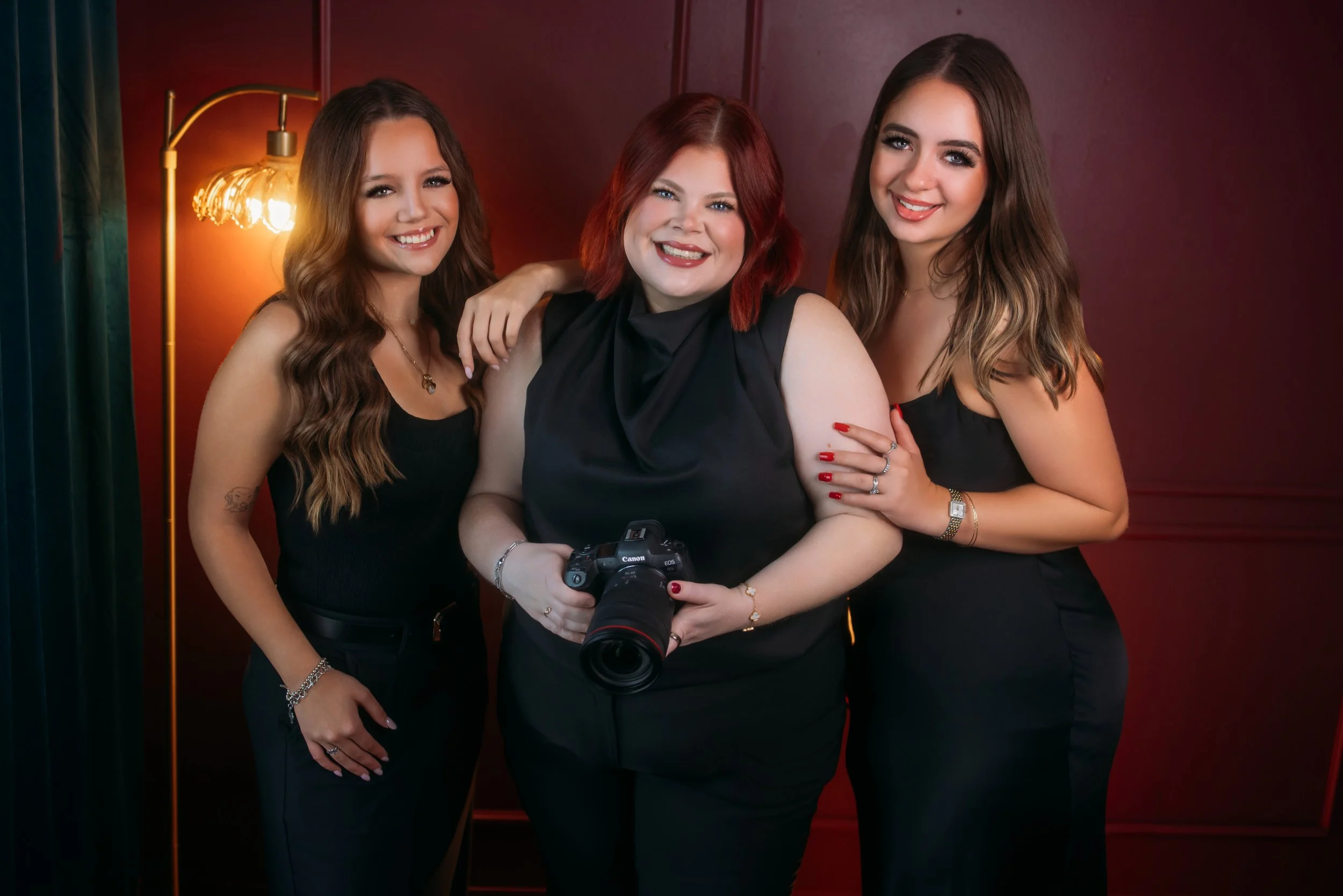 Three women smiling and standing close together, one holding a camera, in front of a red wall with a lit lamp.