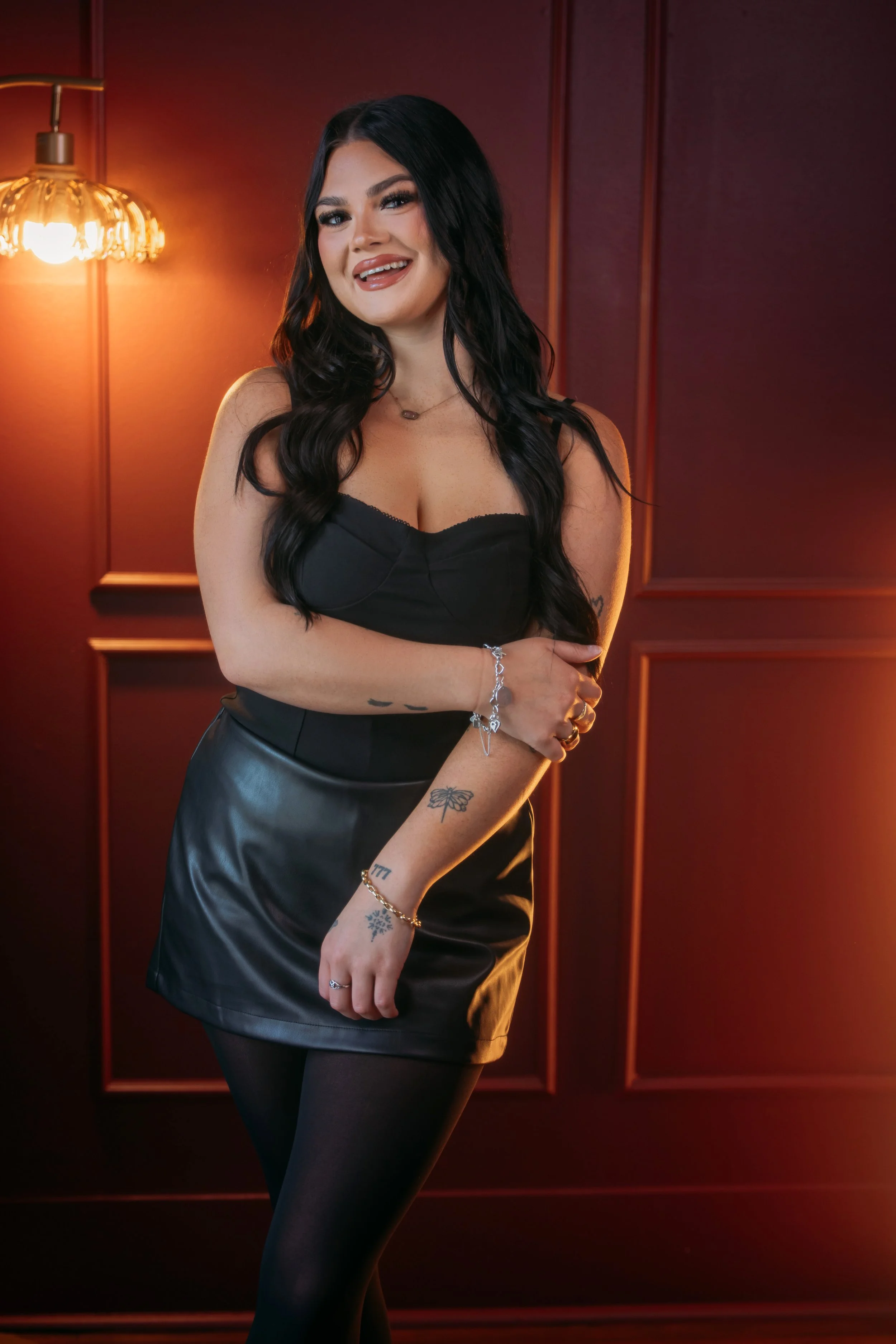 A woman with long black hair and tattoos on her arm, wearing a black strapless top, black skirt, and black tights, smiling and posing with her arms crossed in front of a maroon wall with wood paneling and a vintage brass wall sconce.