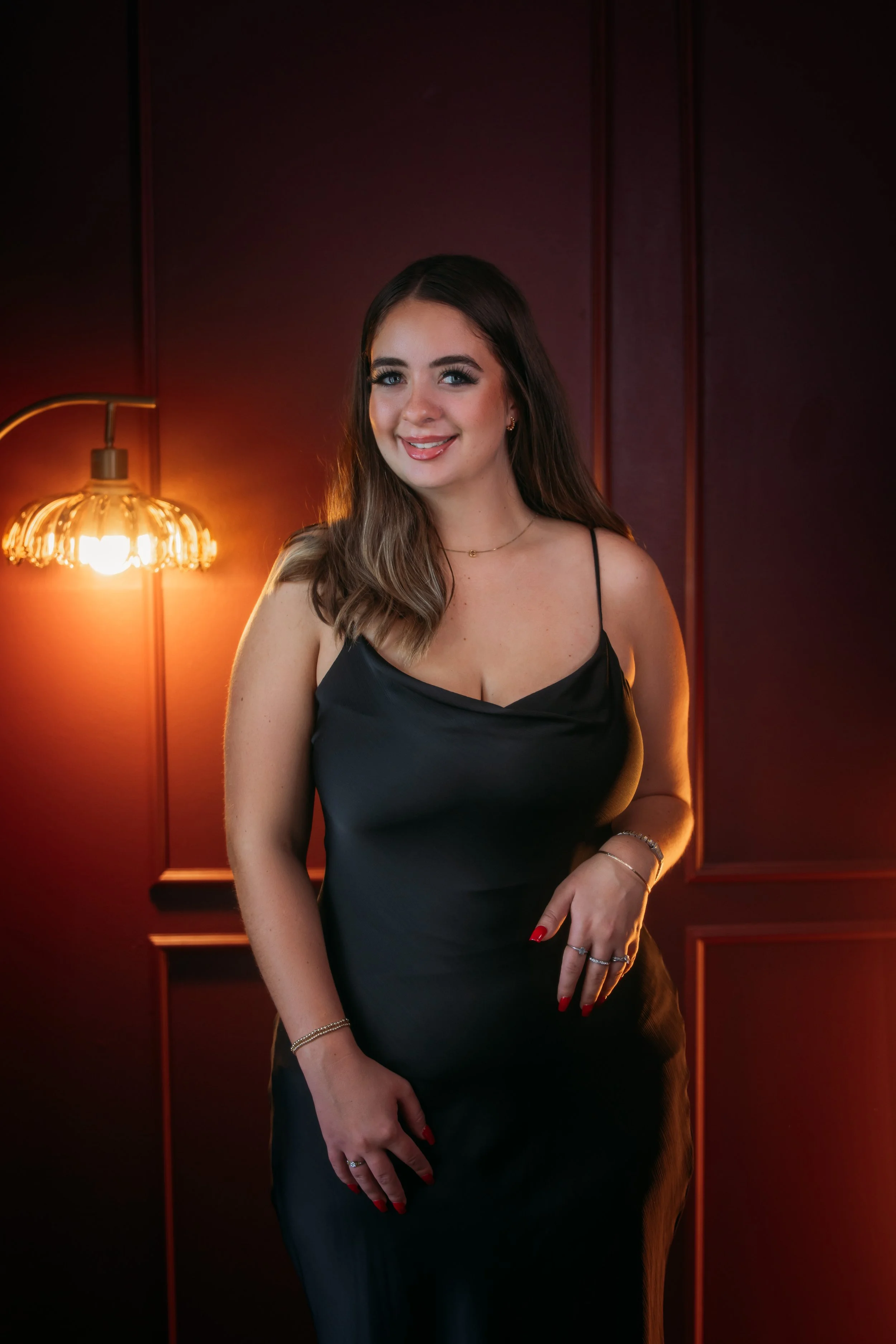 Young woman with long brown hair, blue eyes, wearing a black spaghetti strap dress, standing in front of a dark red wall with wooden panels, illuminated by a warm vintage-style wall lamp.