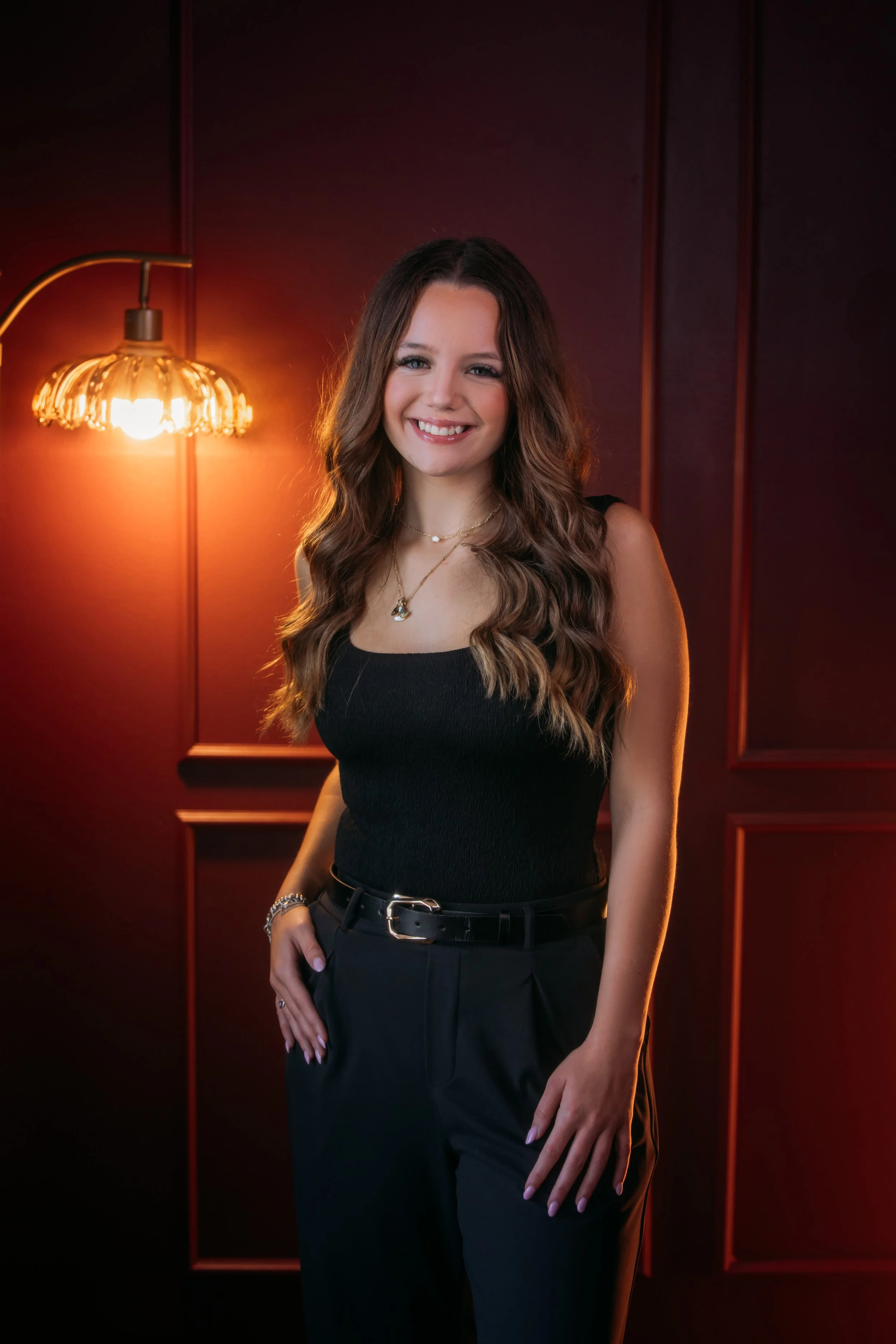 A young woman with long wavy brown hair, wearing a black sleeveless top and black pants, standing in front of a dark red wall with wooden paneling. She is smiling at the camera.