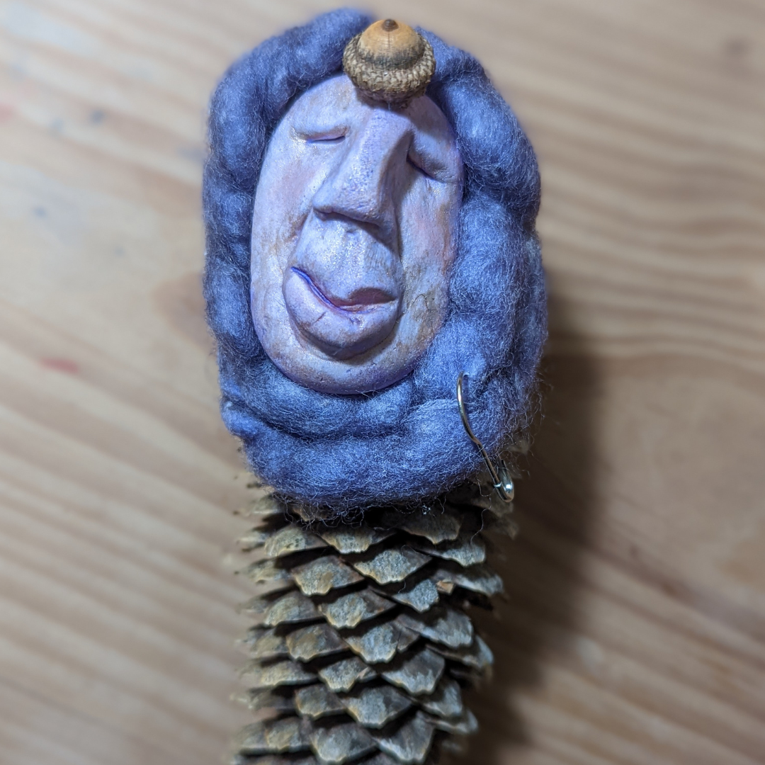 Purple handmade face framed with felted wool and attached to a pinecone doll.