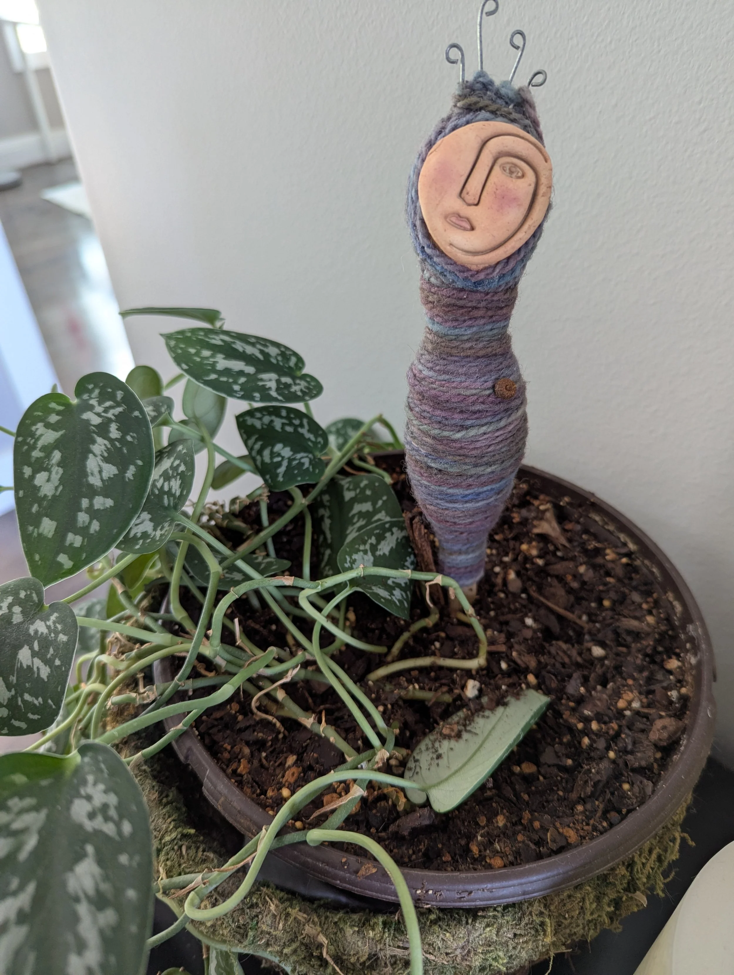 Small handmade plant person with a smiling clay face wrapped in purple yarn, placed in a white pot with a green houseplant