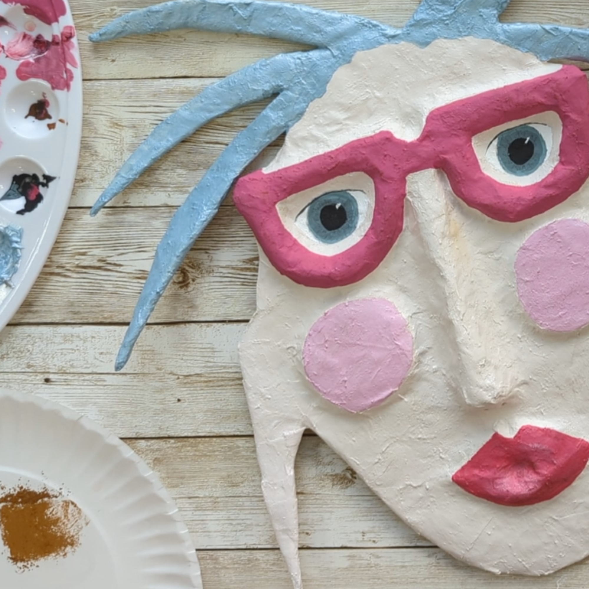 Close-up of a hand-painted spirit mask with blue hair, pink glasses, and expressive eyes on a wooden surface.
