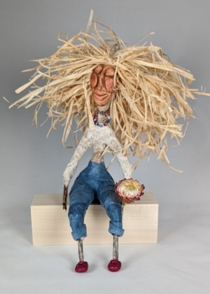 Whimsical spirit doll with straw hair sitting on a wooden block.