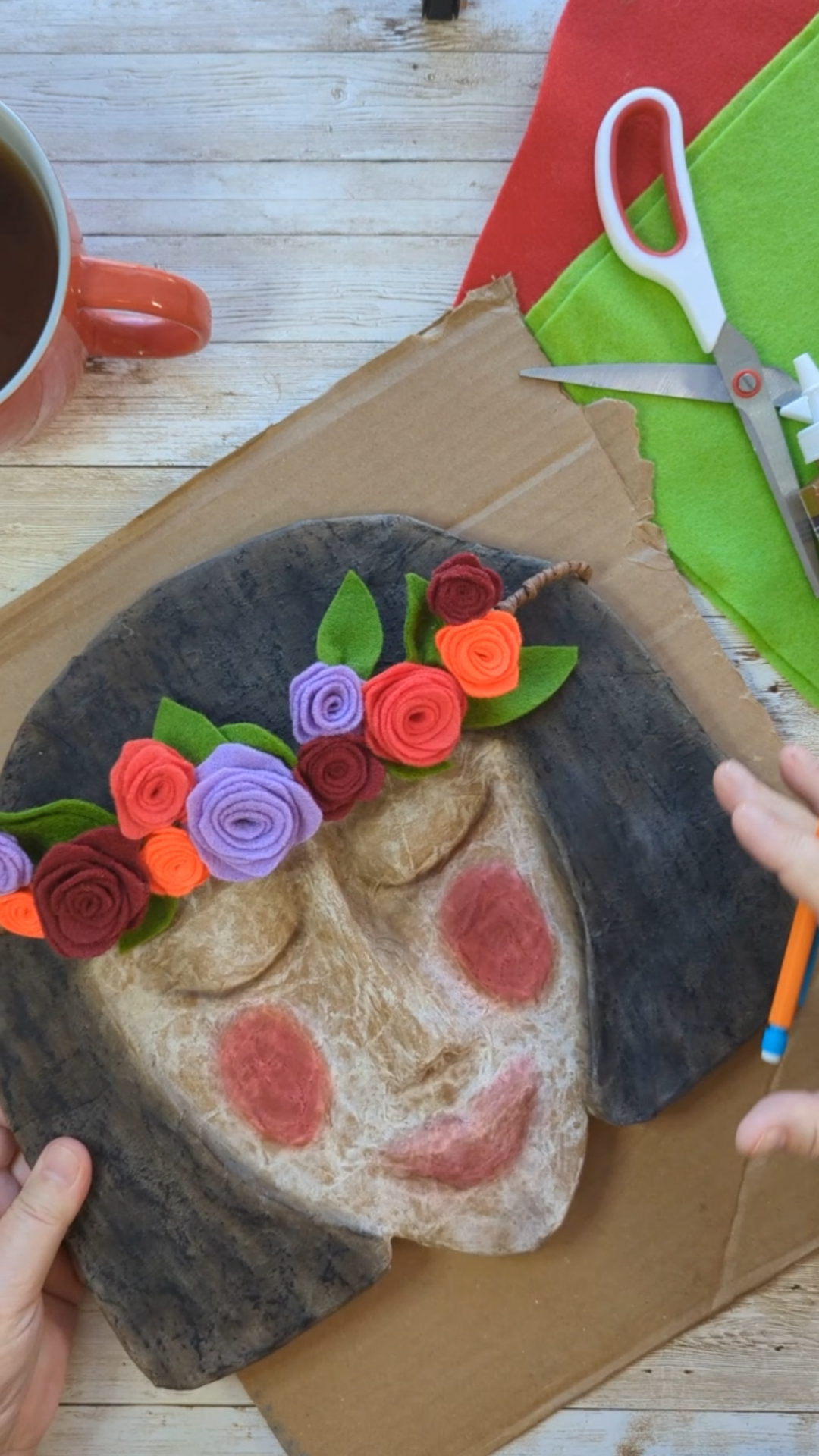 Hands adding colorful felt flowers to a handmade Spirit mask on a rustic table with tea and scissors.