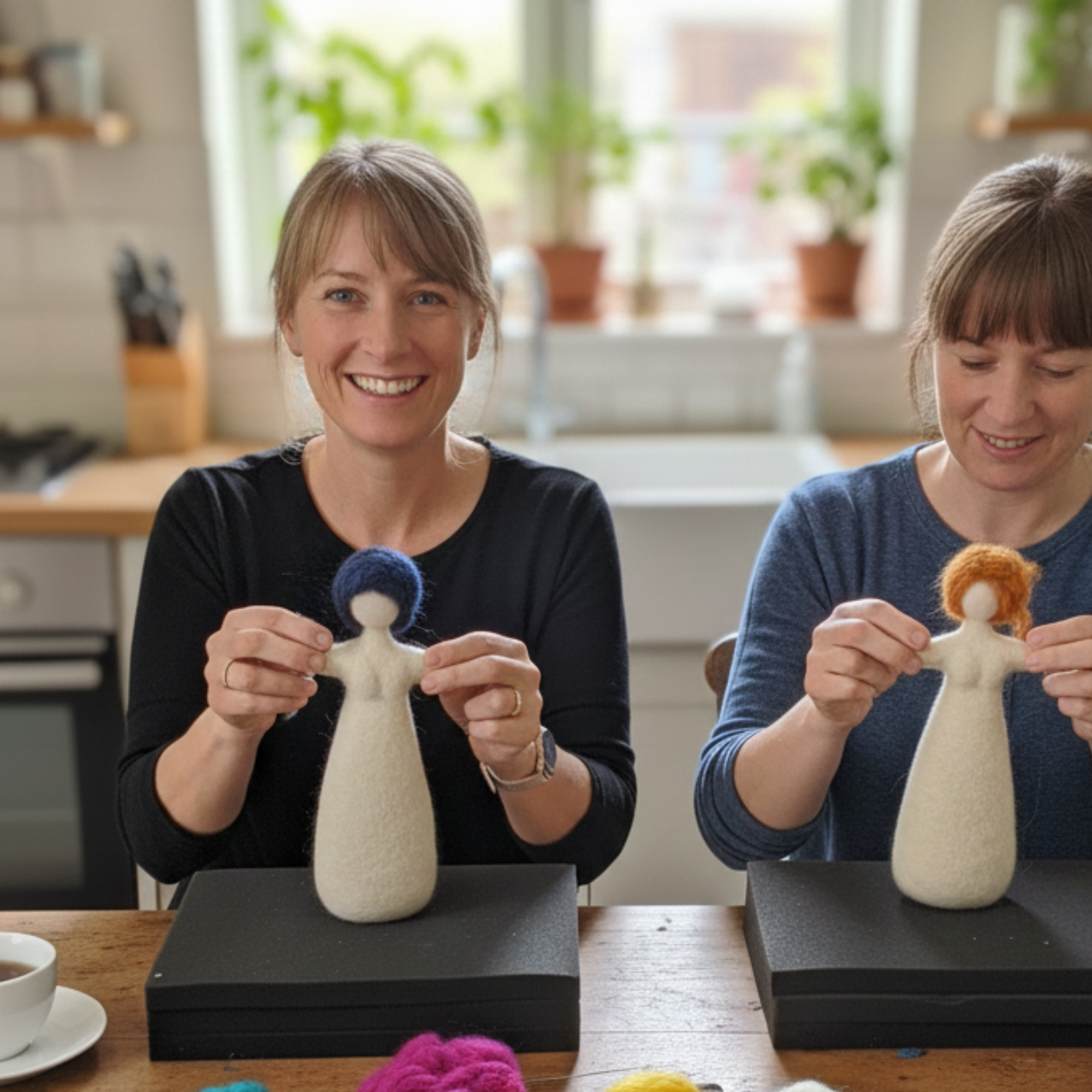 Close-up of two women working side by side during a doll-making retreat, stitching and shaping wool fiber figures with tea on the table.