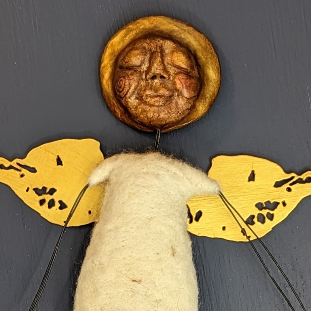 Handmade clay face attached to a small felted angel with golden wings.