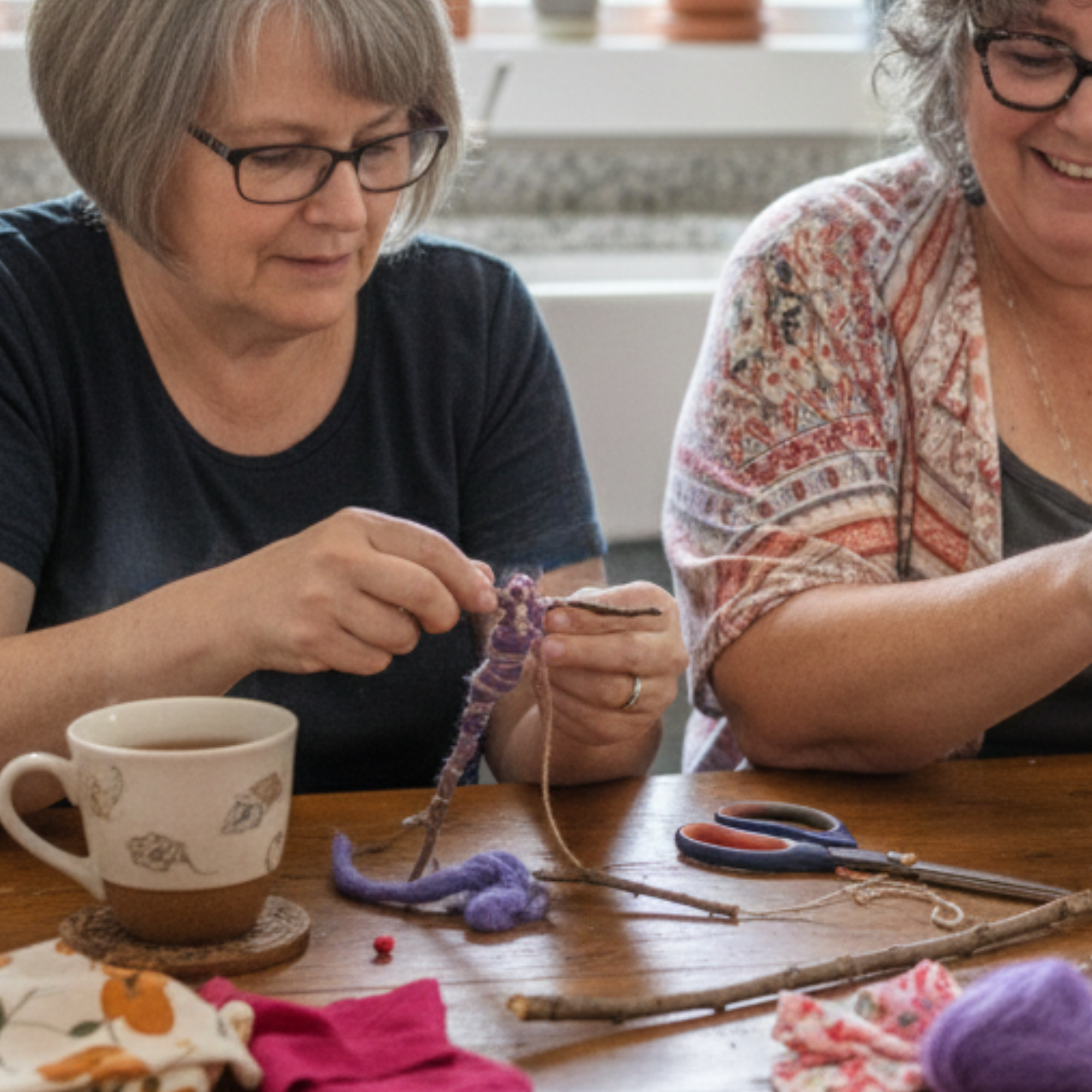 Close-up of two women working side by side during a doll-making retreat, stitching and shaping wool fiber figures with tea on the table.