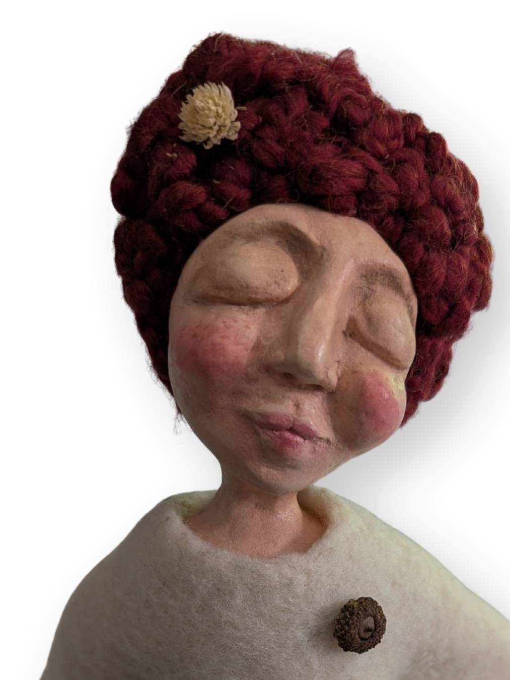 Close-up of a handmade spirit doll with closed eyes, rosy cheeks, deep red yarn hair, and a small acorn attached to her cream-colored clothing.