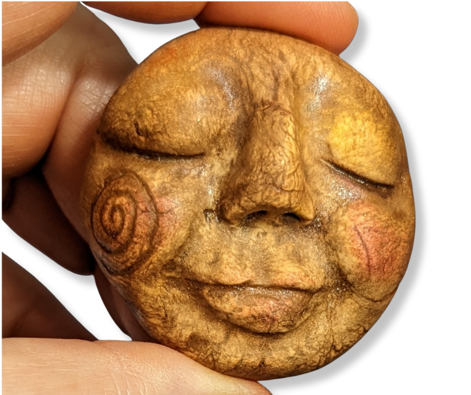 A warm brown clay face for a spirit doll, decorated with spiral designs and held delicately in someone’s fingers—ready to be brought to life.