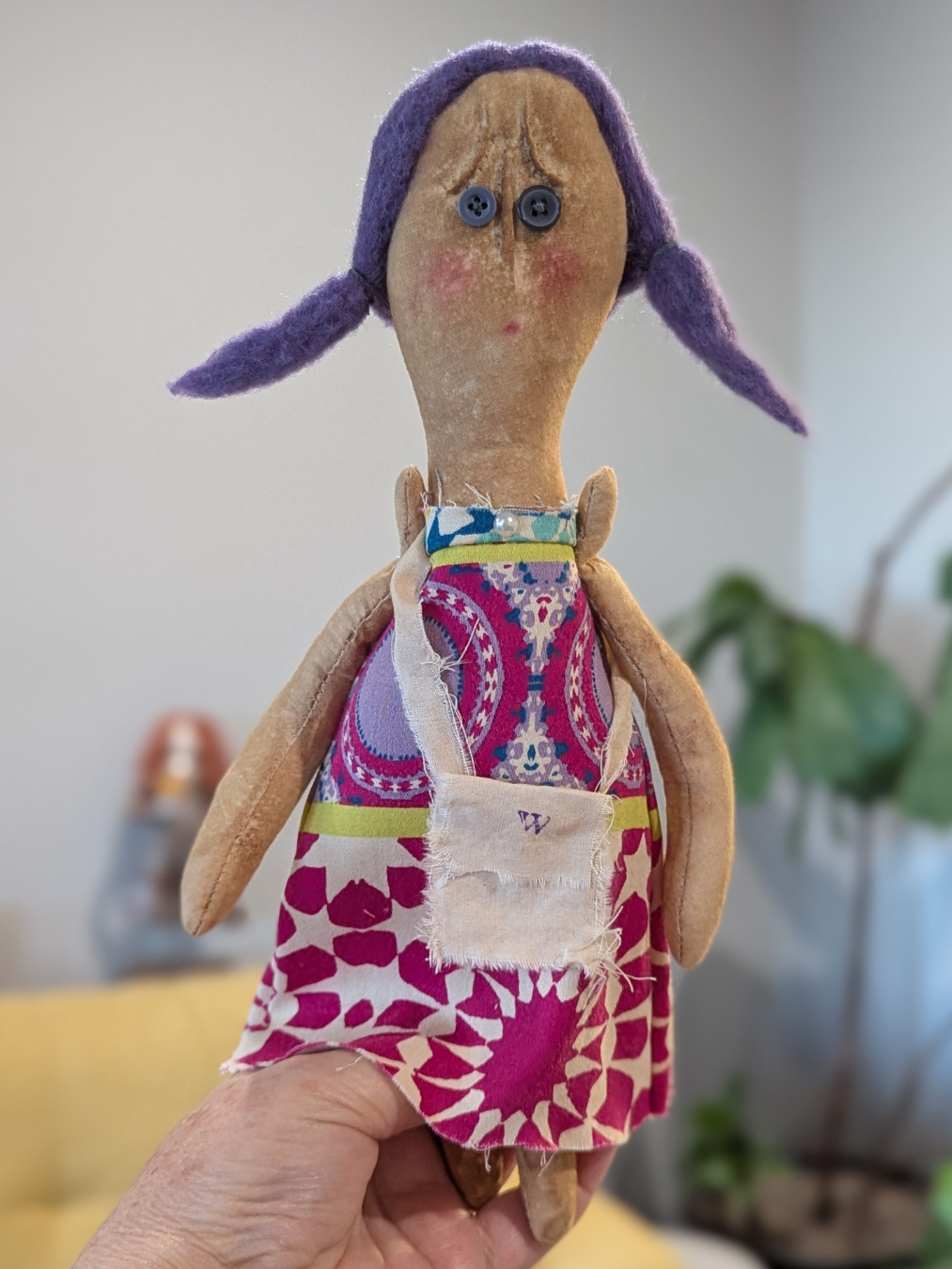 Handmade worry doll with purple wool pigtails, button eyes, and a pink and puple dress held up for display.