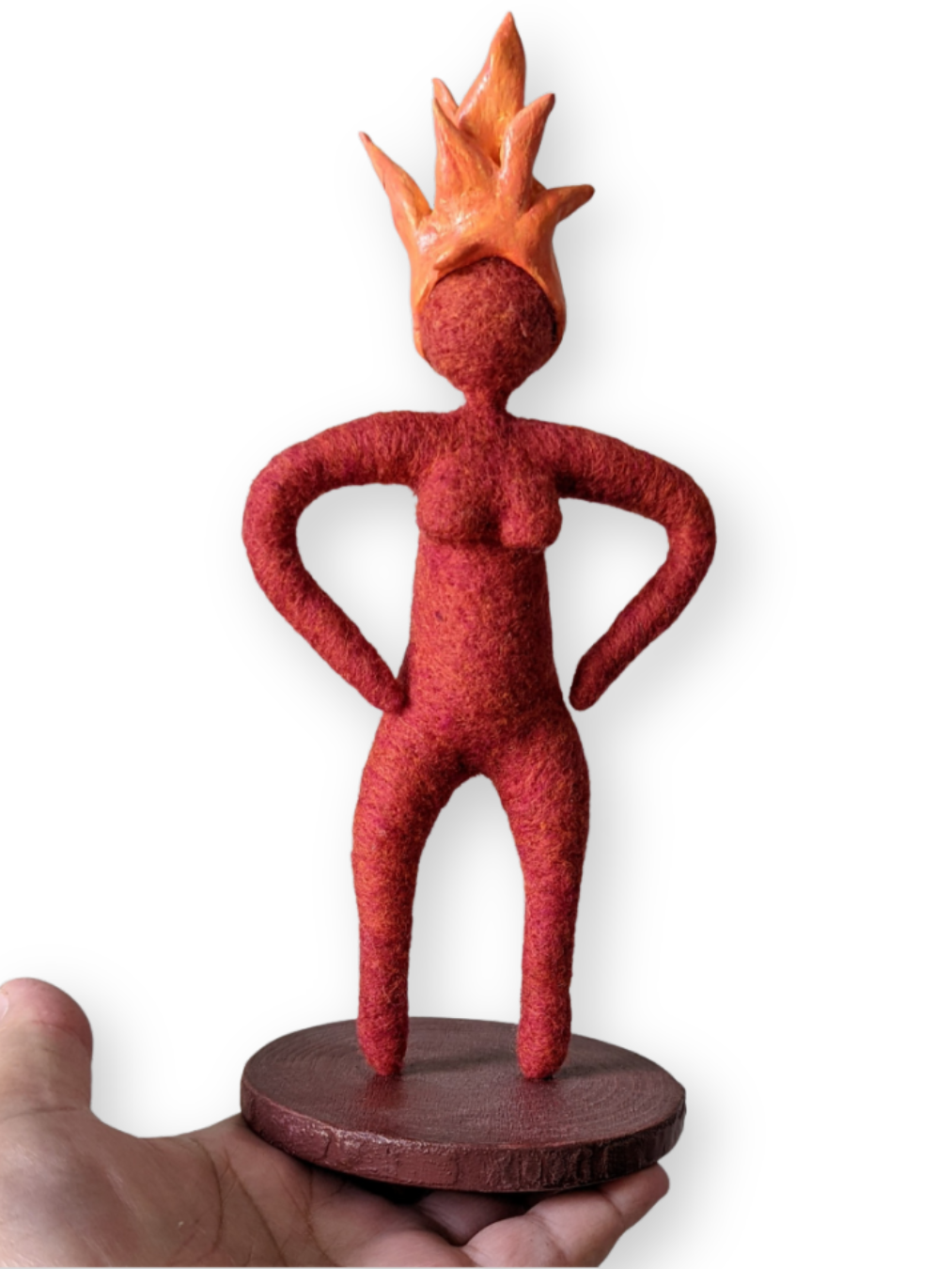 A small handmade needle-felted spirit doll in deep red wool with a bright orange flame-shaped crown, standing confidently with hands on hips atop a round wooden base, symbolizing inner fire and strength.