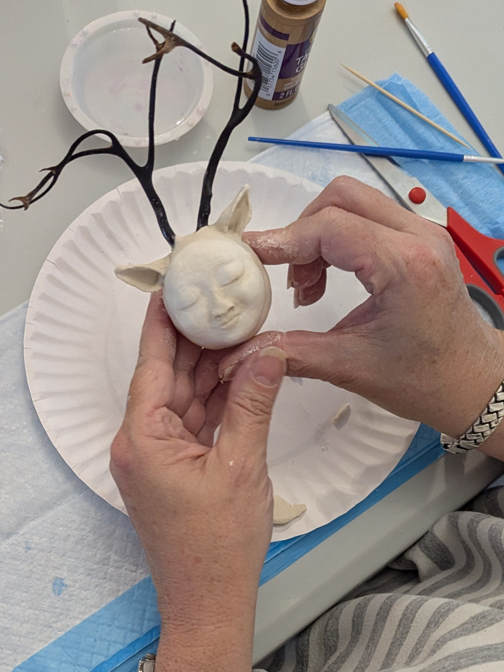 Hands shaping a clay face with small ears for a stick doll made from branches.