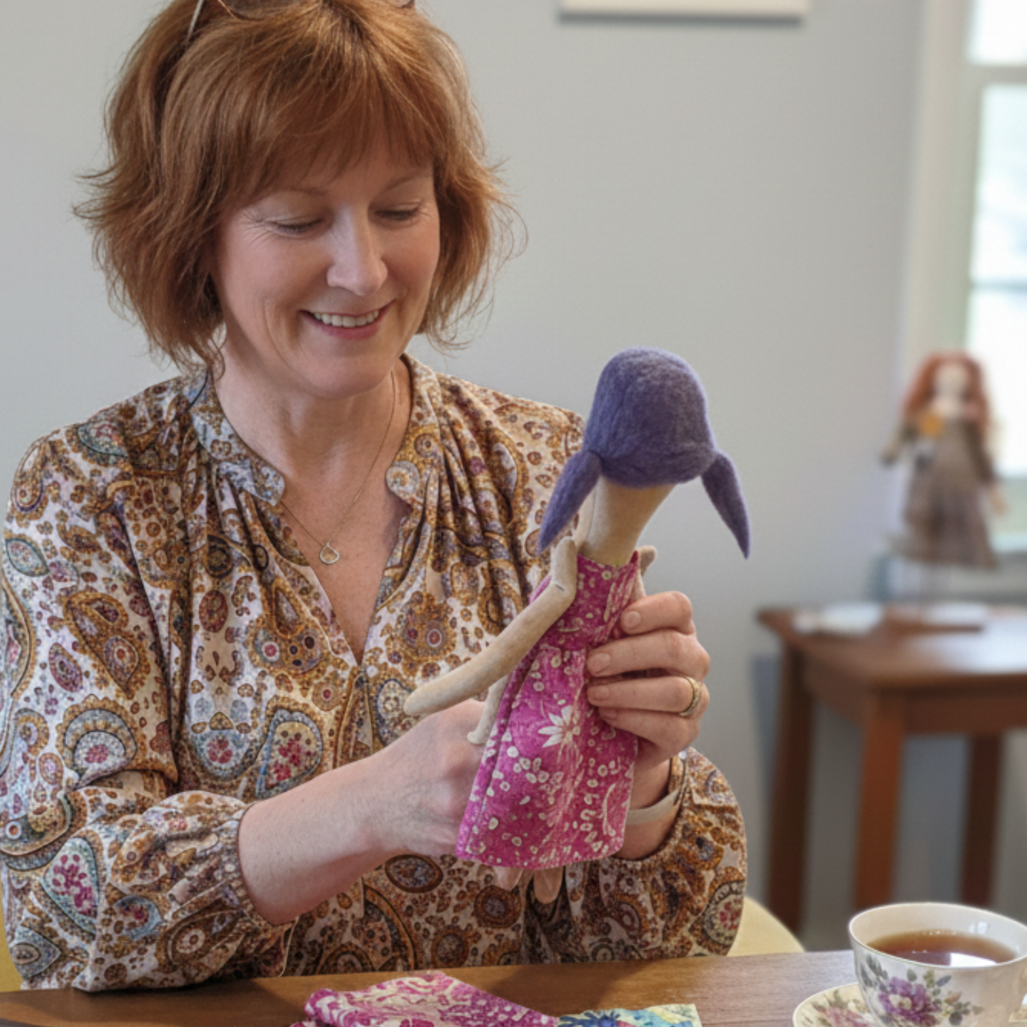 Woman smiling at the fabric worry doll she made during the workshop, seated at a table with tea.