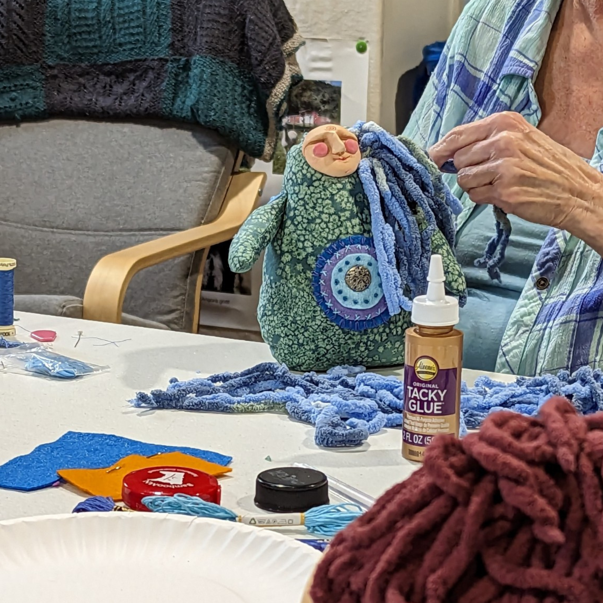 Hands adding yarn hair to a green poppet doll during a live workshop, surrounded by fabric, thread, and supplies.