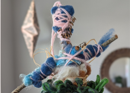 Small fiber talisman wrapped in blue and pink wool roving, tied with thread and dried botanicals, resting on a branch as part of the Quickening Bundle project.