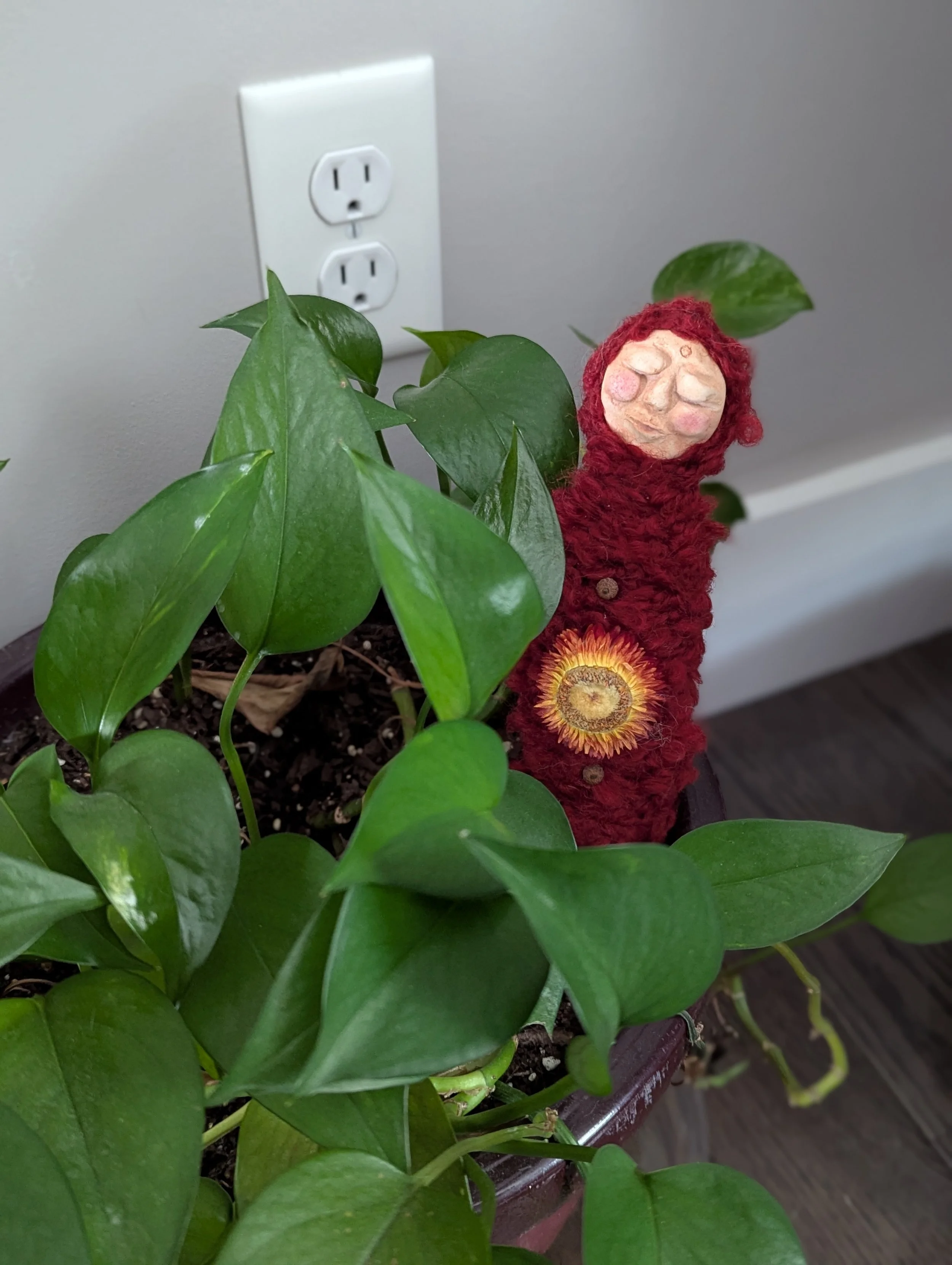 Handmade plant person with a clay face wrapped in red yarn, decorated with a small flower, nestled in a leafy potted houseplant